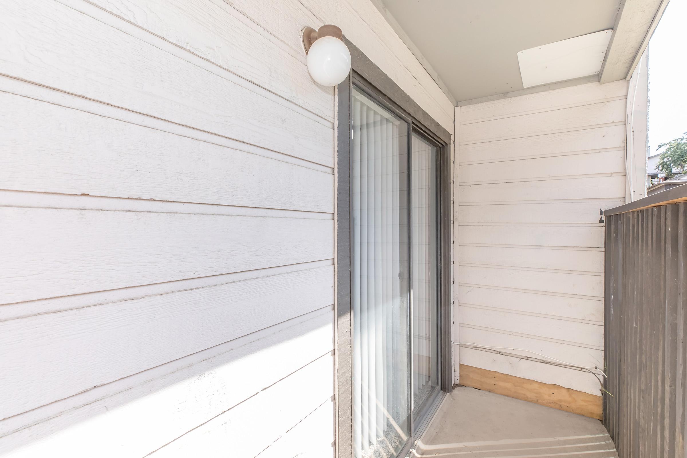 A corner of a porch with white wooden siding and a glass sliding door. There's a light fixture mounted above and visible floor space. Sunlight casts soft shadows on the exterior surfaces.