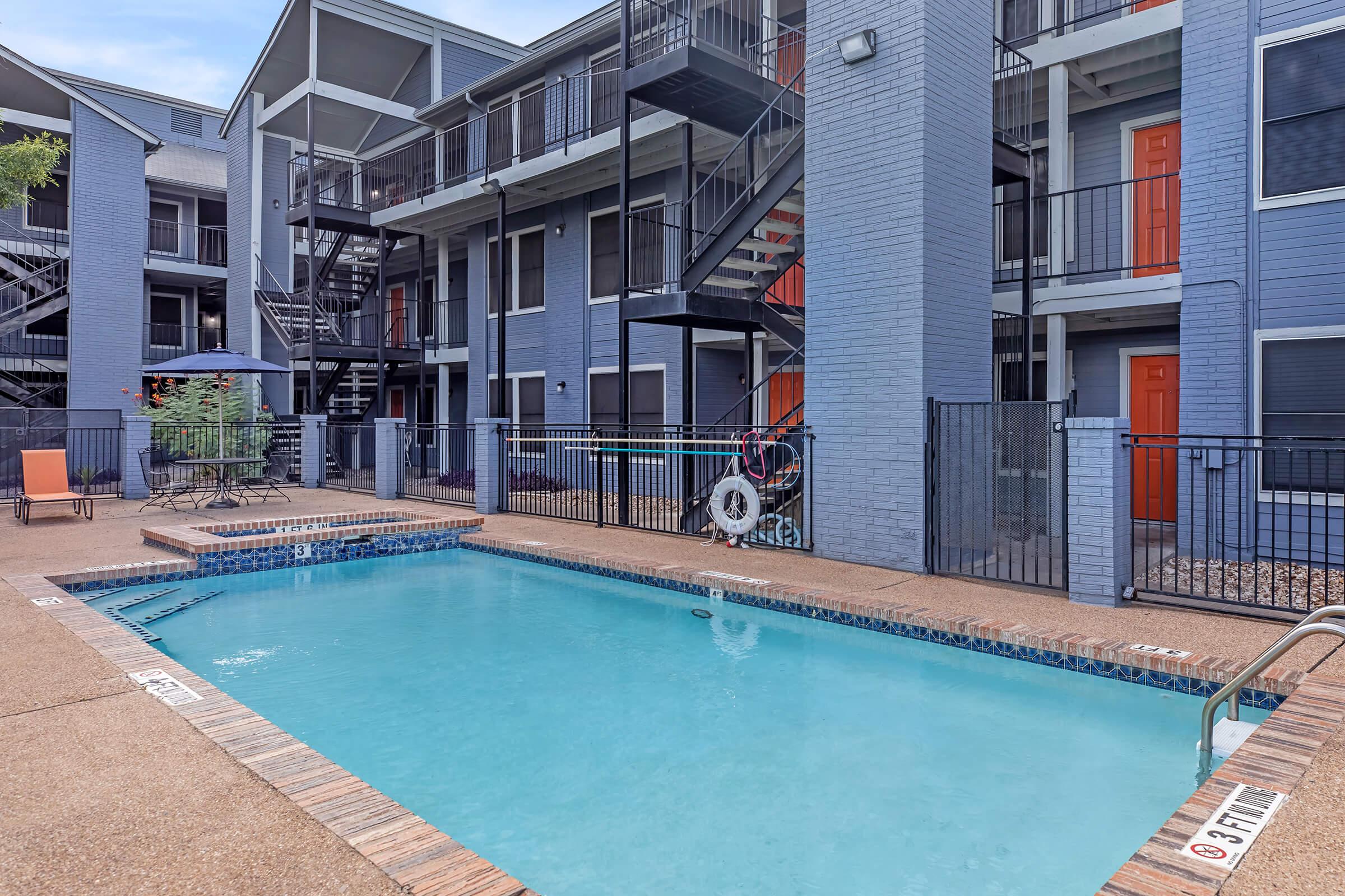 A view of an apartment complex featuring a swimming pool, lounge chairs, and surrounding dark blue buildings with orange doors. There are metal railings and an outdoor seating area, with stairs leading to upper floors in the background.