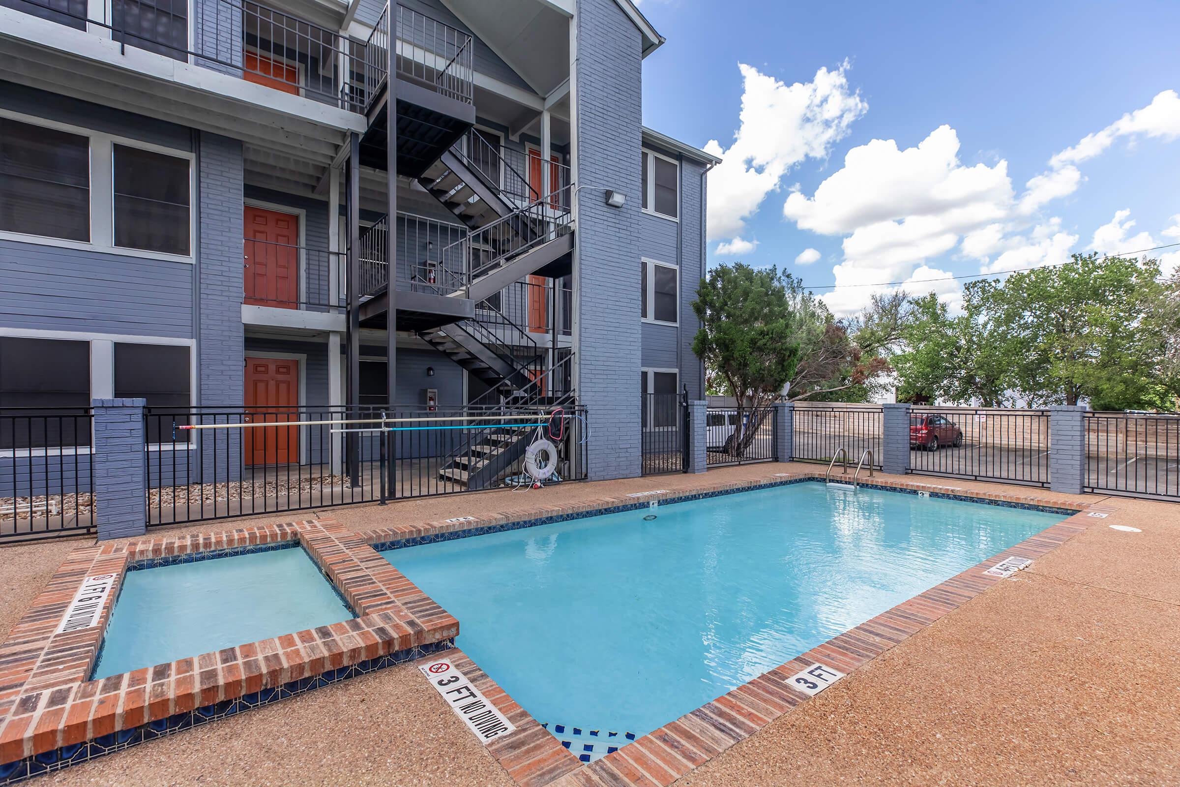 A view of a swimming pool surrounded by a tan patio area. The pool features brick edges and depth markers. Behind the pool, there is a three-story apartment building with a staircase and several windows. The sky is partly cloudy, suggesting a sunny day.