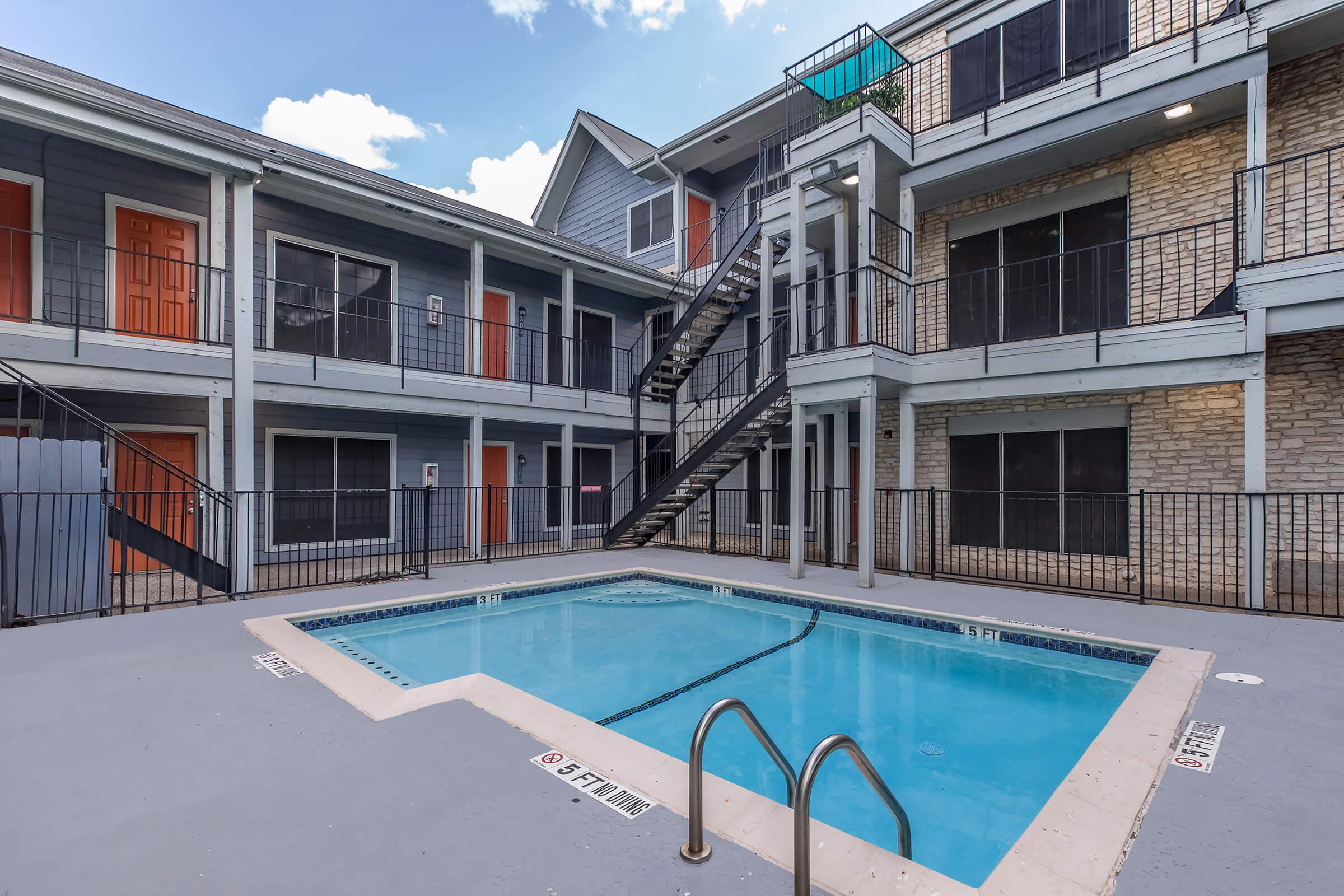 A view of a rectangular swimming pool surrounded by an apartment complex. The pool is located in a courtyard area, with multiple stories of apartments visible in the background. The buildings feature a mix of gray and tan siding, with orange doors and black railings, under a partly cloudy sky.