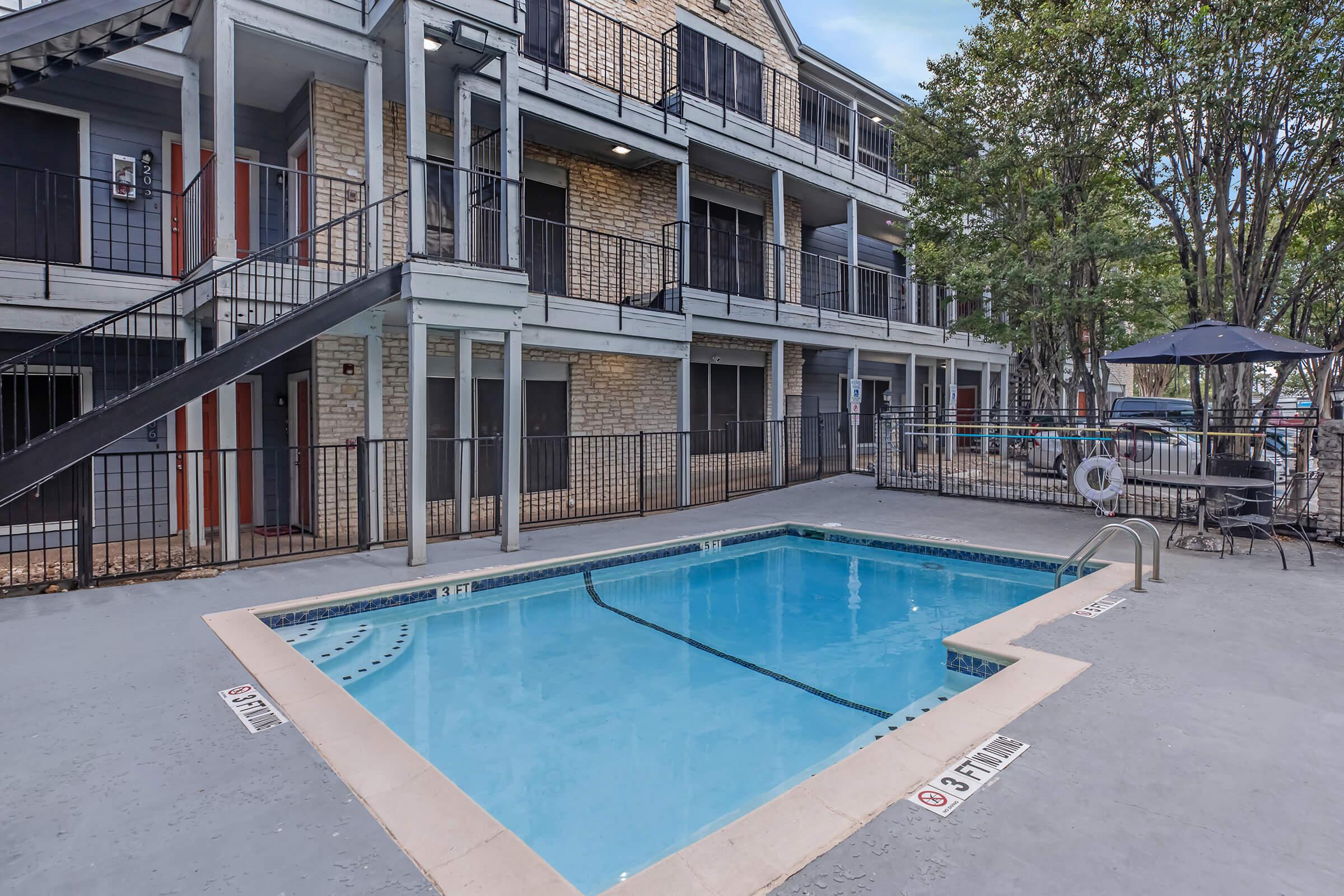 A view of a small outdoor swimming pool surrounded by a concrete deck, adjacent to a multi-story apartment building with balconies. The area features a black metal railing and a sun umbrella near the pool. Signage indicates the pool is marked for depth on two sides.