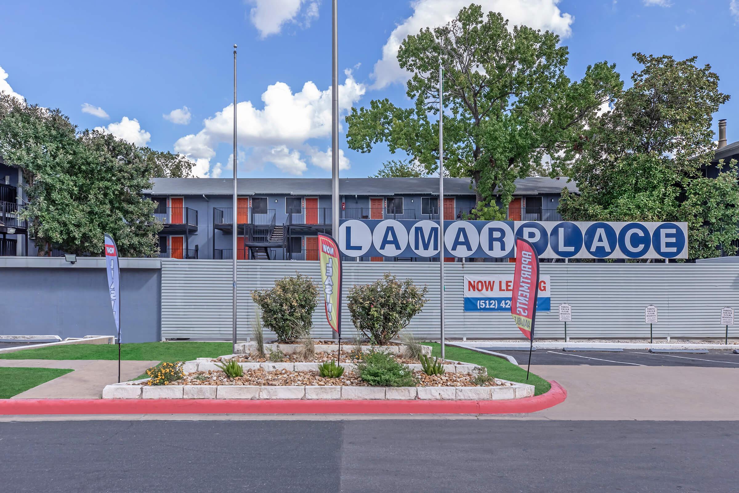 A large sign reading "LAMAR PLACE" at an apartment complex with a landscaped area in the foreground. The entrance features several flags displaying "NOW LEASING" messages. The building has multiple balconies and is surrounded by trees under a partly cloudy sky.