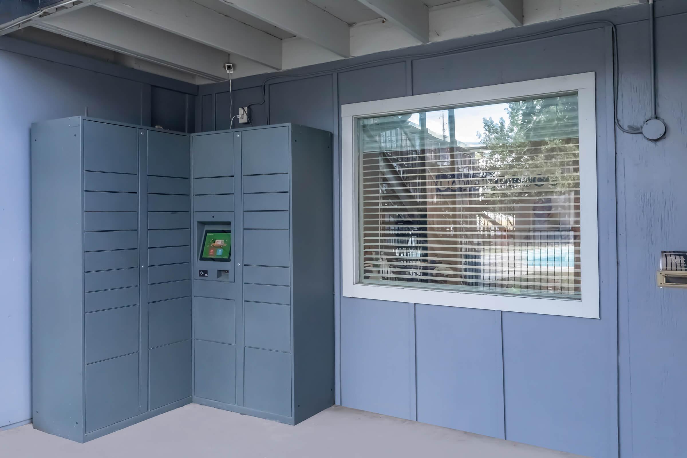 A gray delivery locker system is positioned next to a window with blinds. The locker has a digital interface on the front, and the wall is painted light blue. There is a slightly shaded area outside near a pool, which can be seen through the window.