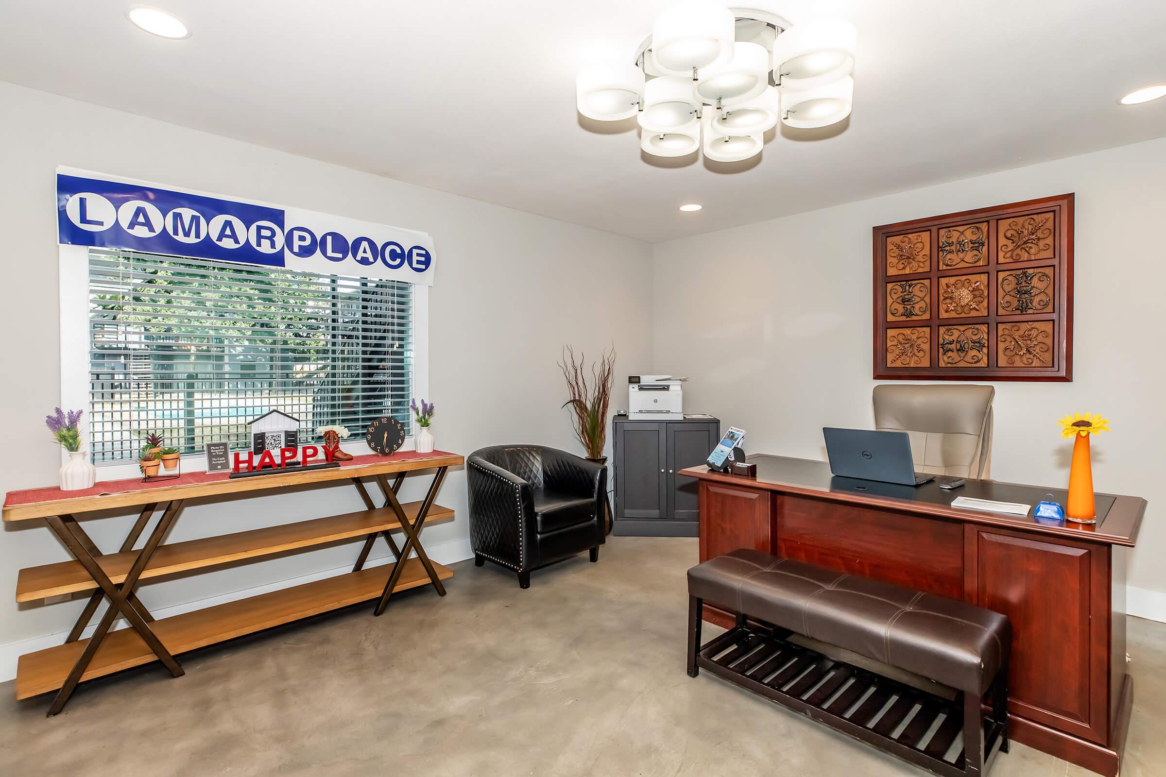 A modern office interior featuring a desk with a computer, a decorative wooden table with a small house model, and a dark chair. Natural light enters through a window with curtains. A stylish light fixture hangs from the ceiling, and decorative elements like plants are present. The wall has a framed art piece.