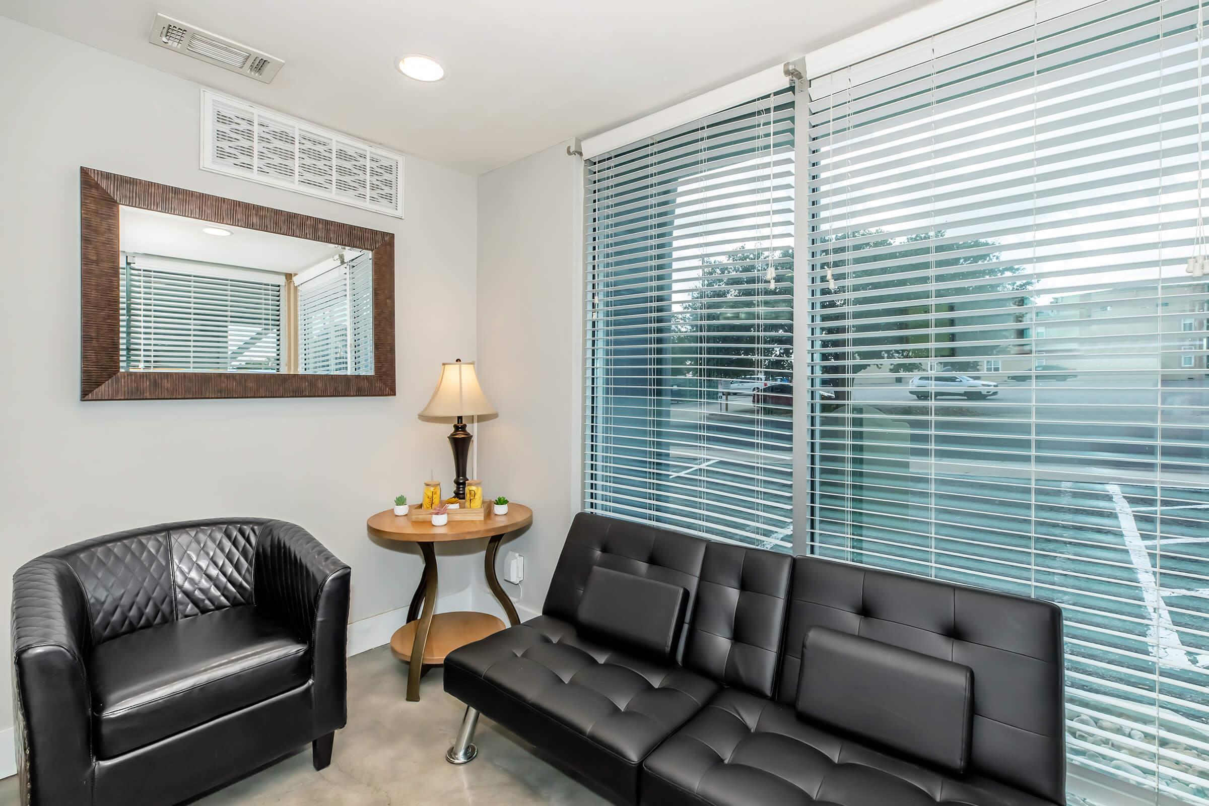 A modern waiting area featuring a black leather sofa and an armchair. There's a round side table with decorative items, a lamp, and a large mirror on the wall. Natural light filters through the blinds on a window, creating a bright and inviting atmosphere.