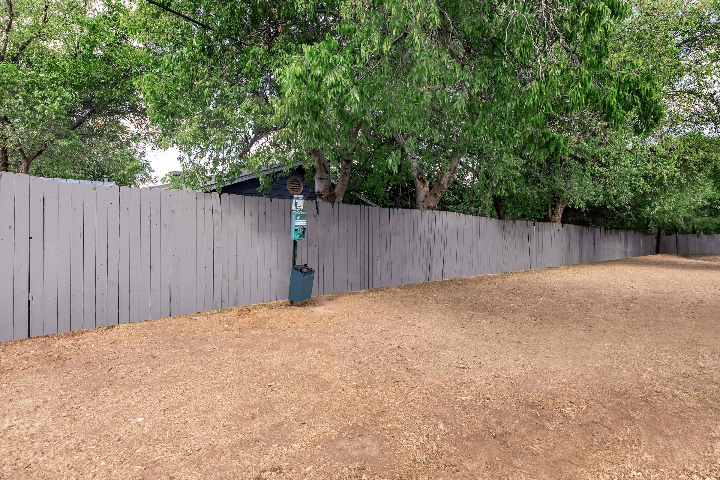 A gray wooden fence runs along a dirt path, with green trees providing shade on both sides. A blue trash bin is positioned near the fence, and a small sign or mailbox is visible attached to the fence. The area appears quiet and natural, with no people or buildings in sight.