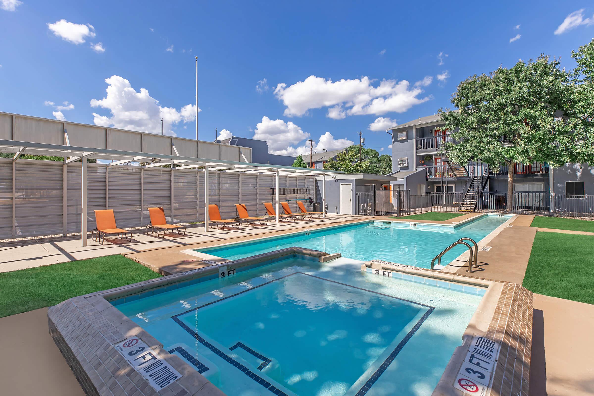 Crystal-clear swimming pool with attached hot tub, surrounded by lounge chairs under shade structures. Bright blue sky with fluffy clouds overhead, and a multi-story apartment building in the background. Lush green grass enhances the area around the pool.