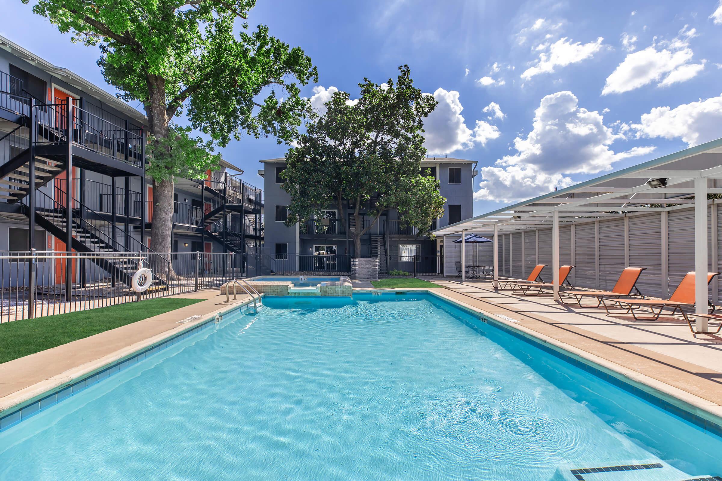A sunny outdoor pool area featuring a clear blue swimming pool surrounded by lounge chairs. In the background, there are multi-story buildings with balconies and staircases, along with lush trees providing shade. Bright blue skies with scattered clouds complete the inviting atmosphere.