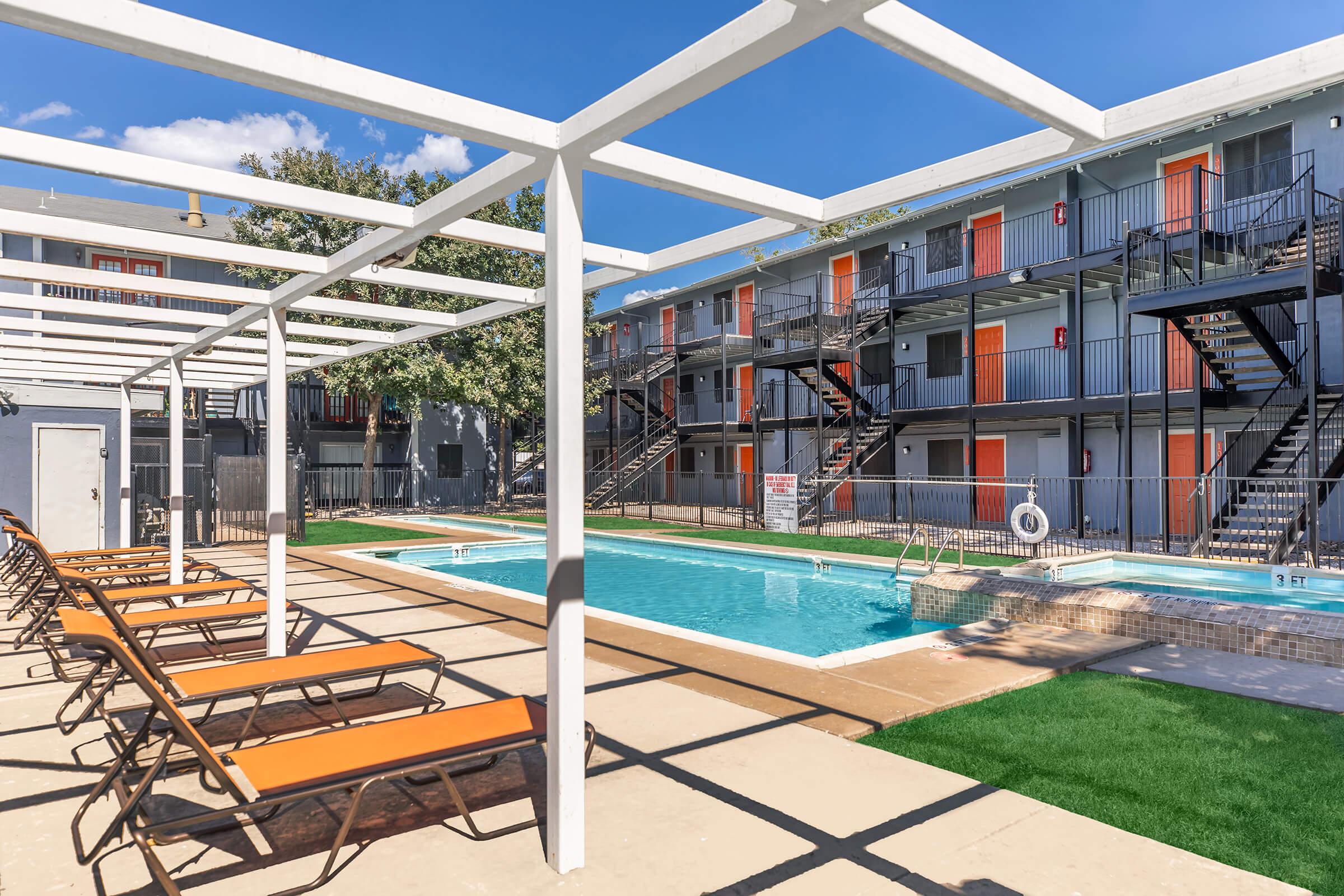 A view of a pool area surrounded by lounge chairs. The pool is clear and reflects the blue sky, with two orange doors visible on a building in the background. There are white pergolas overhead, and a grassy area nearby, creating a relaxing atmosphere for visitors.