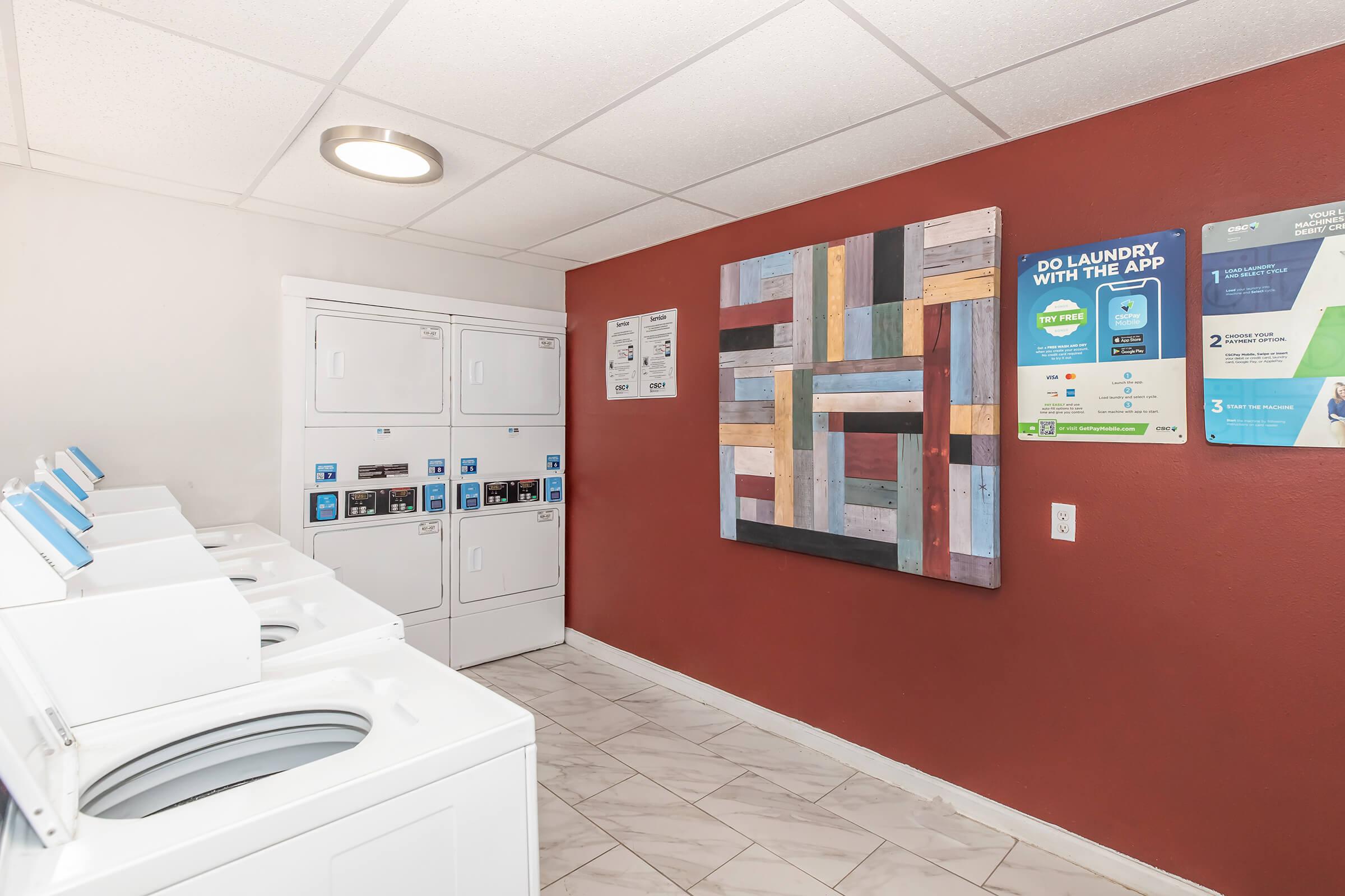 A brightly lit laundry room featuring several white washing machines and dryers against a red accent wall. There’s a colorful wooden art piece on the wall and informational posters about laundry app services. The floor is tiled in light colors, contributing to a clean and modern atmosphere.