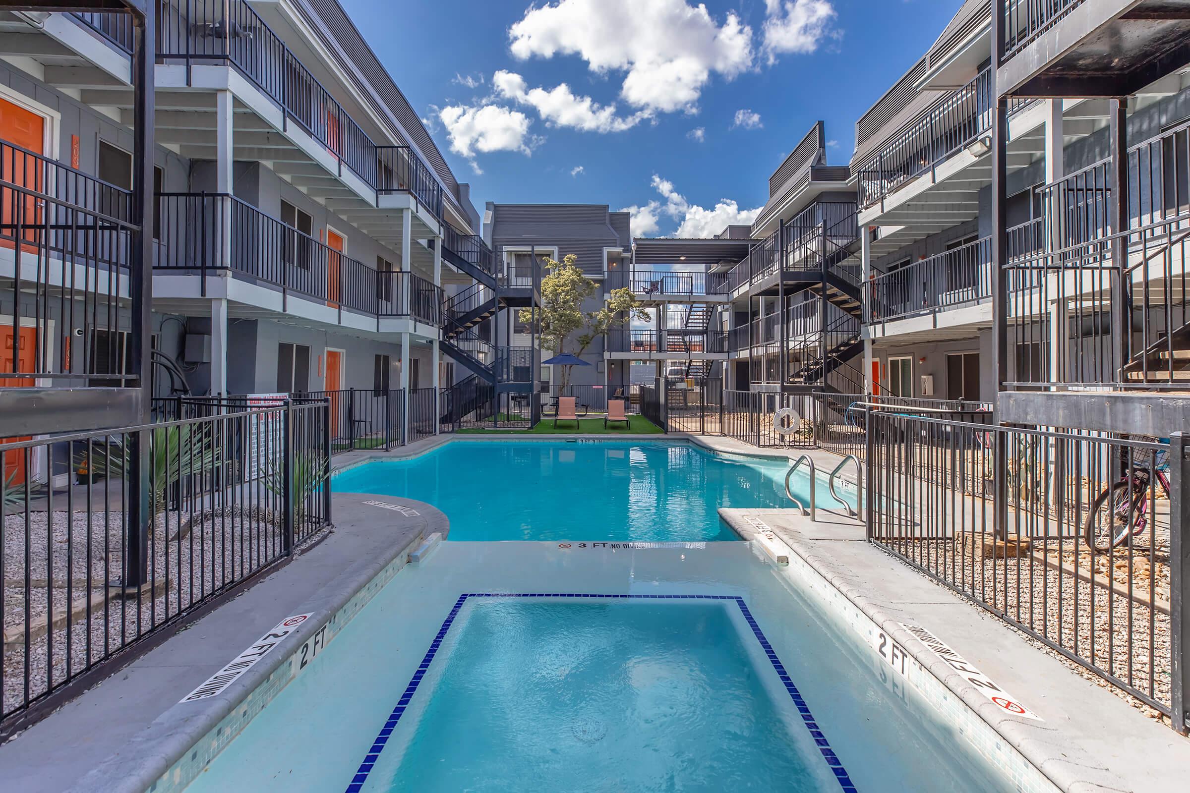 A view of a modern apartment complex featuring two-story buildings surrounding a swimming pool. The pool area has lounge chairs and is framed by palm trees, with bright blue skies and fluffy white clouds in the background. The balconies of the apartments have black railings, adding to the contemporary aesthetic.