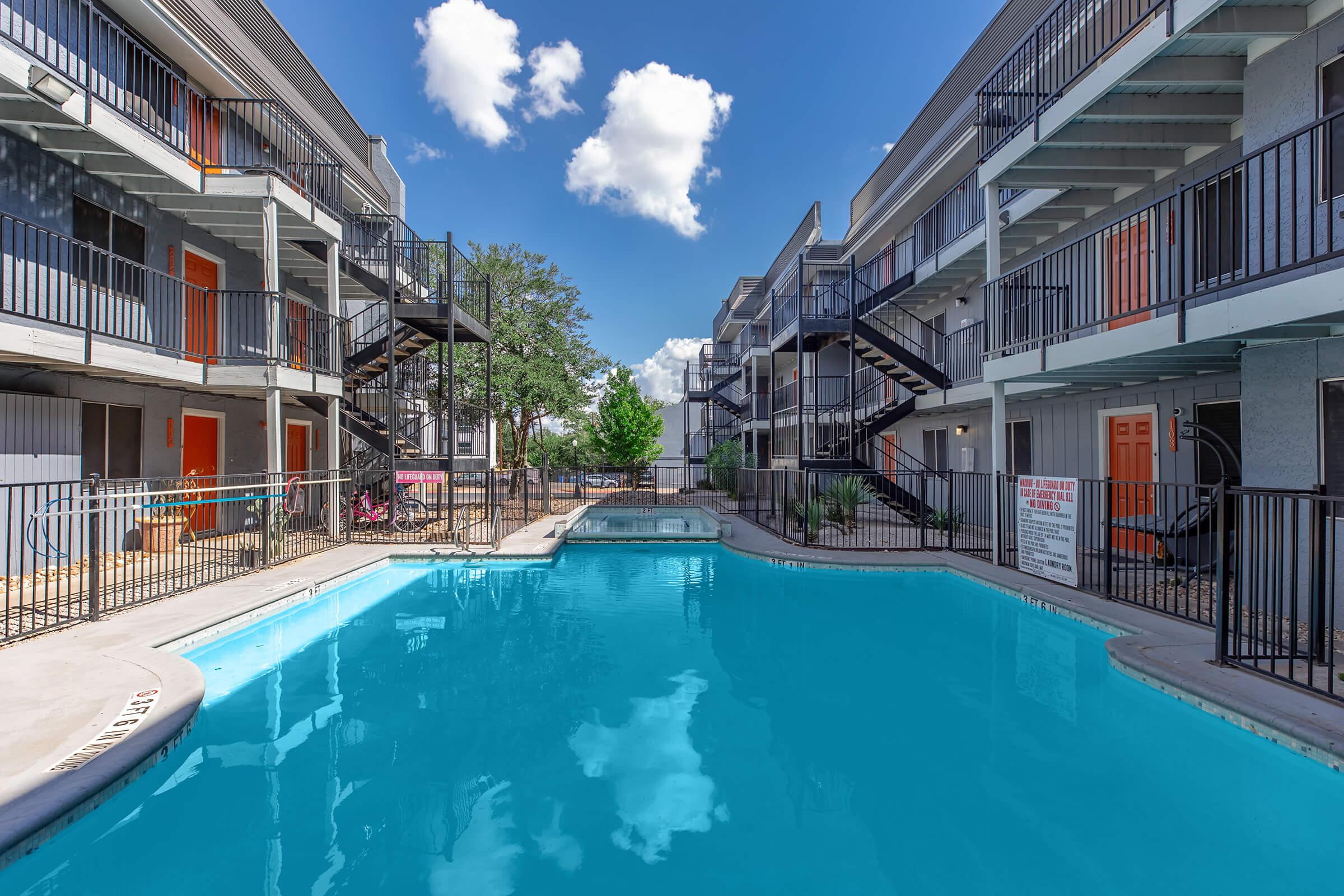 A clear blue swimming pool surrounded by two multi-story apartment buildings with balconies. The buildings have orange doors and black railings, and there are trees and a seating area nearby. The sky is bright with a few fluffy clouds.