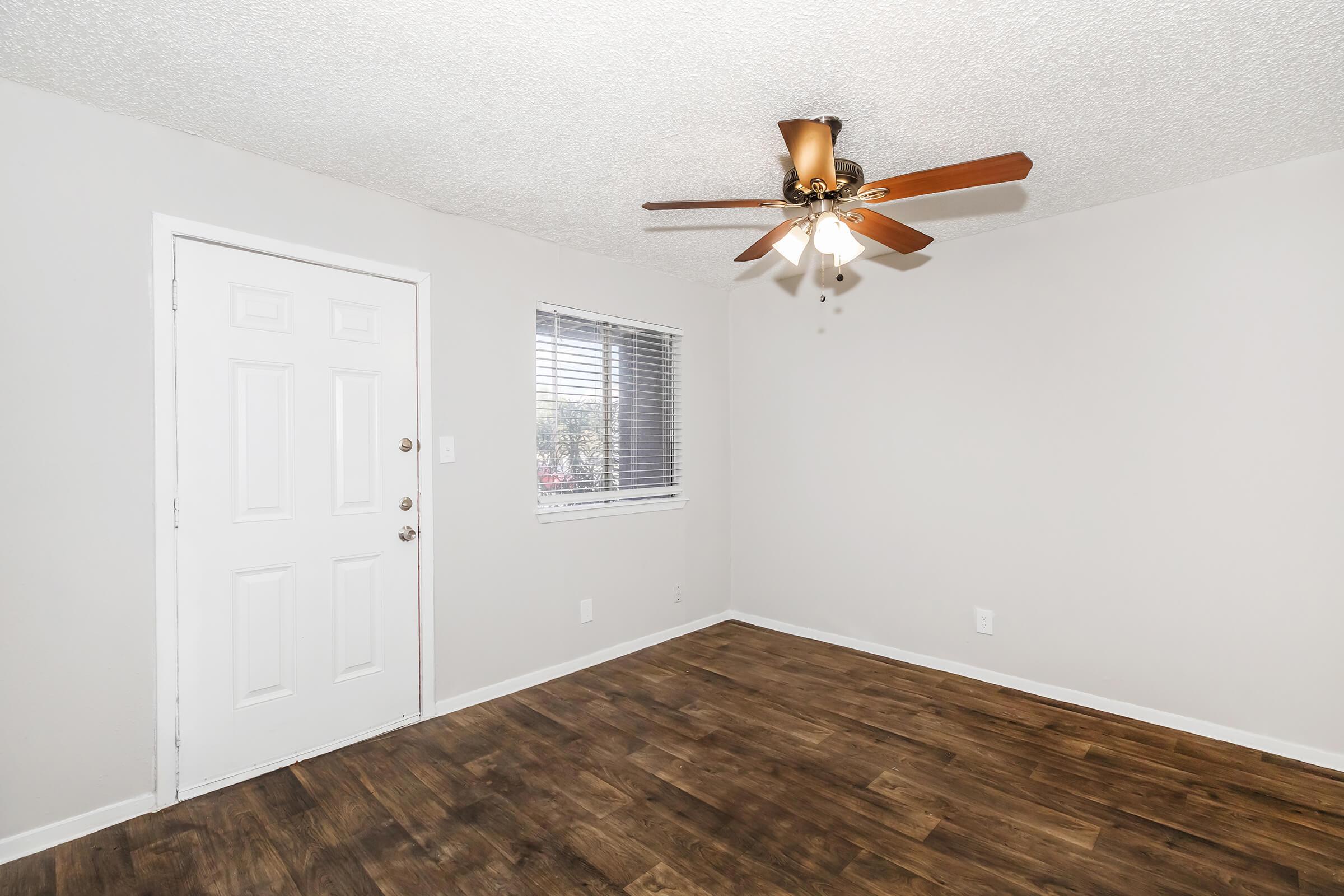 Bright, empty room with a tan ceiling fan, light walls, and wood laminate flooring. A closed white door is on the left, and a window with blinds is on the right, allowing natural light to enter. The room has no furniture, creating a spacious and minimalist appearance.
