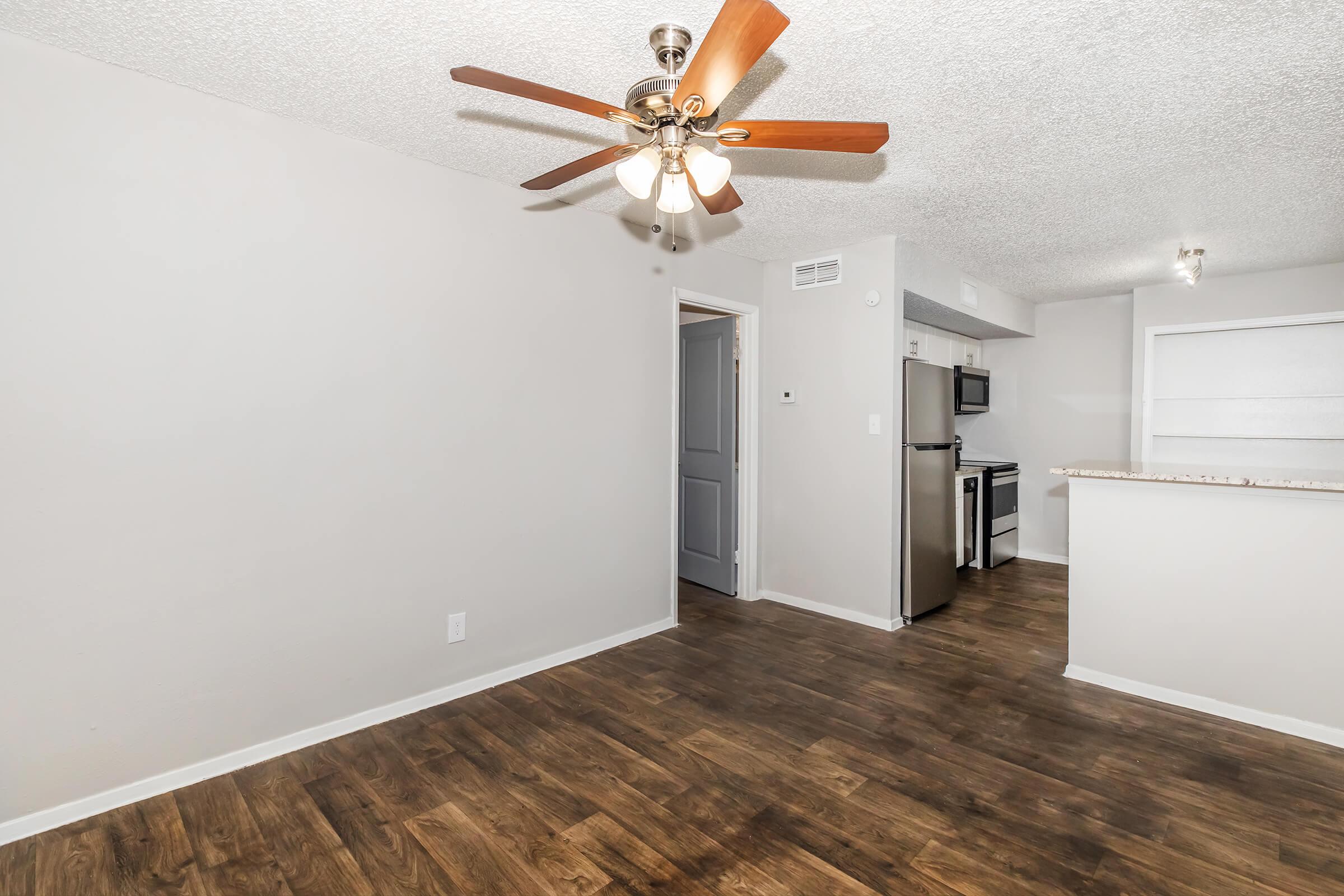 A spacious living area featuring a light-colored ceiling with a ceiling fan, wooden flooring, and neutral-colored walls. There is a doorway leading to another room, and an open kitchen area with stainless steel appliances visible in the background. Natural light brightens the space.