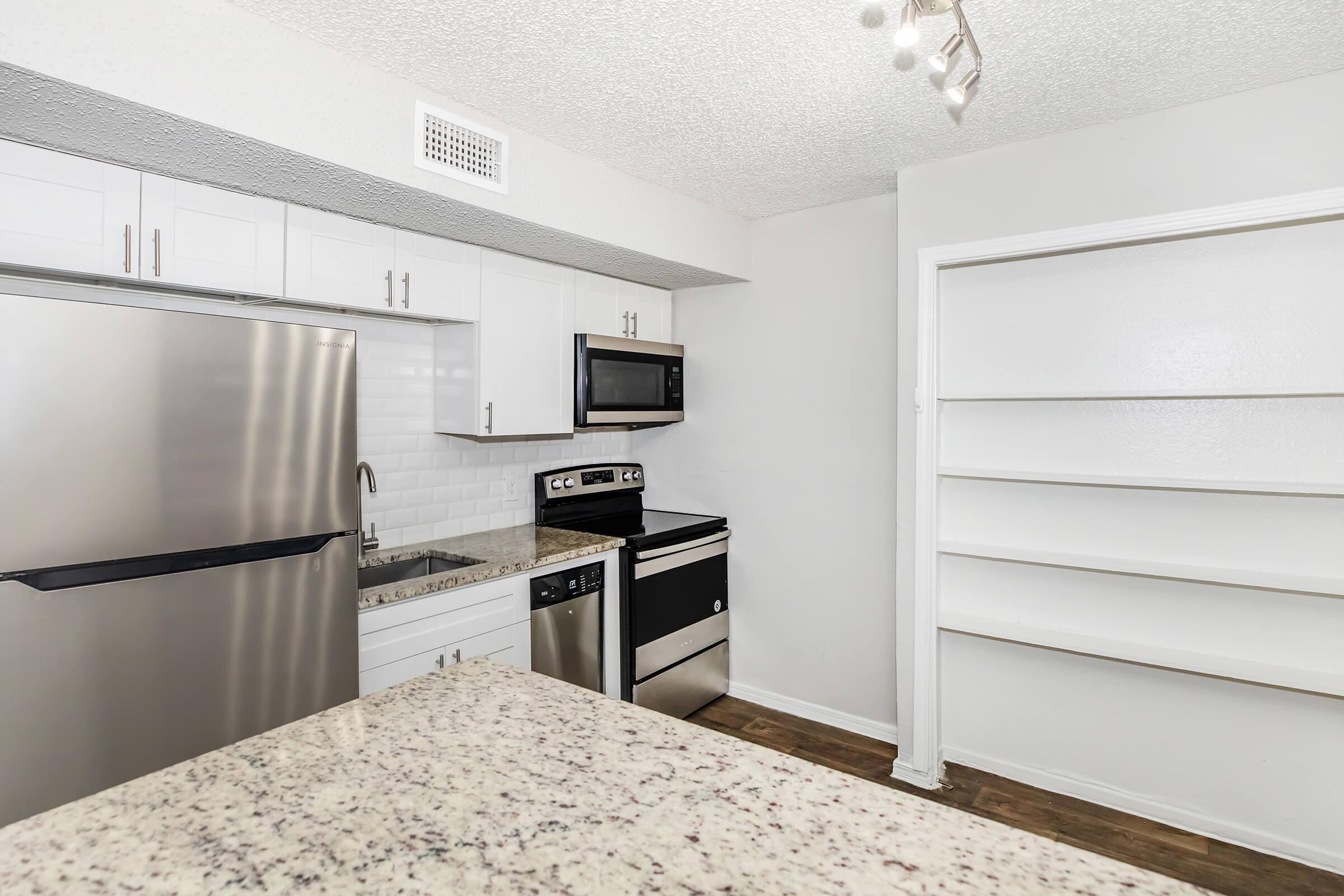 Modern kitchen featuring stainless steel appliances, including a refrigerator, microwave, and oven. White cabinetry and a granite countertop add a sleek touch, while a pantry or storage area is visible on the right. The overall design is bright and clean, with a neutral color palette and wood-like flooring.