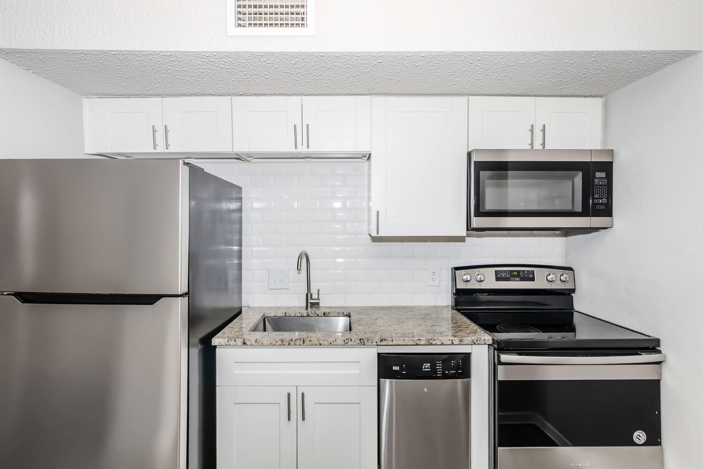 A modern kitchen featuring stainless steel appliances, including a refrigerator, oven, microwave, and dishwasher. The cabinetry is white with clean lines, and the countertops are granite. The backsplash consists of small white tiles, creating a bright and contemporary look.