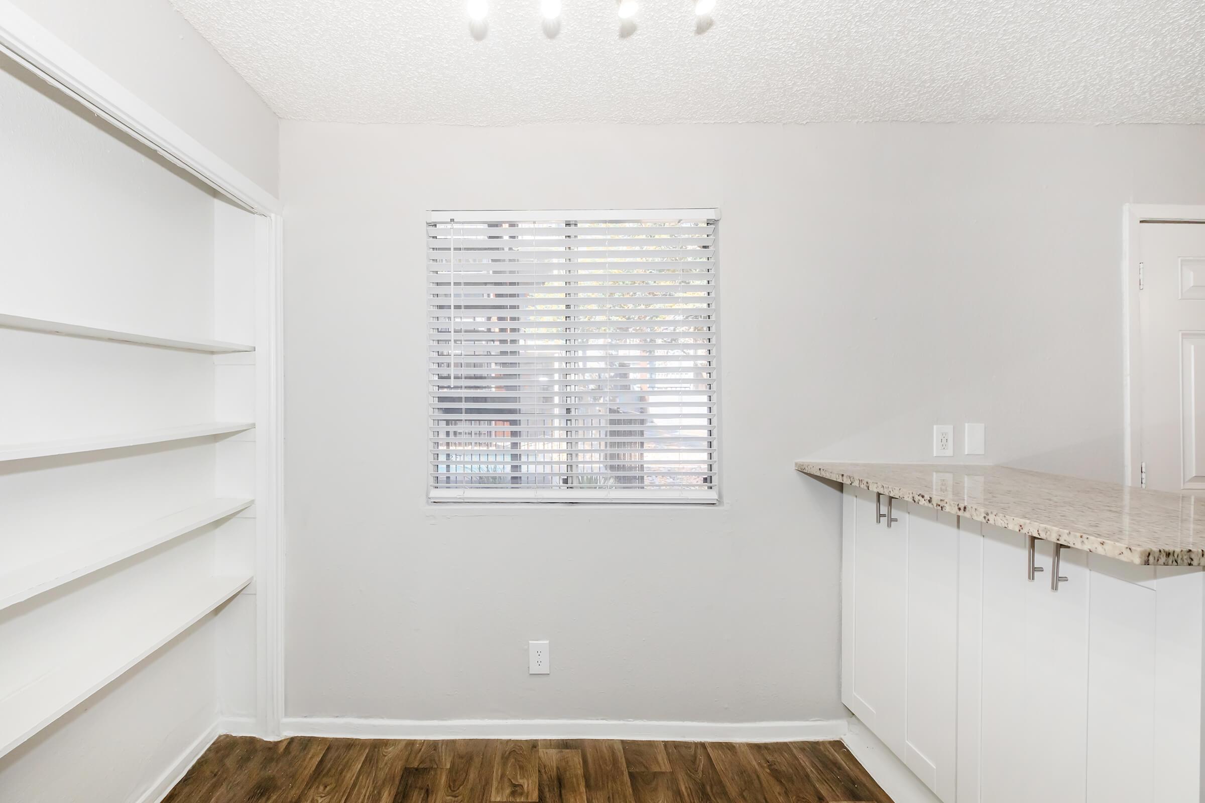 A bright and airy interior space featuring a window with blinds, a light-colored wall, built-in shelves on one side, and a modern bar countertop with a light-colored finish. The floor is made of wood, adding warmth to the room. Ideal for a cozy living area or kitchen.