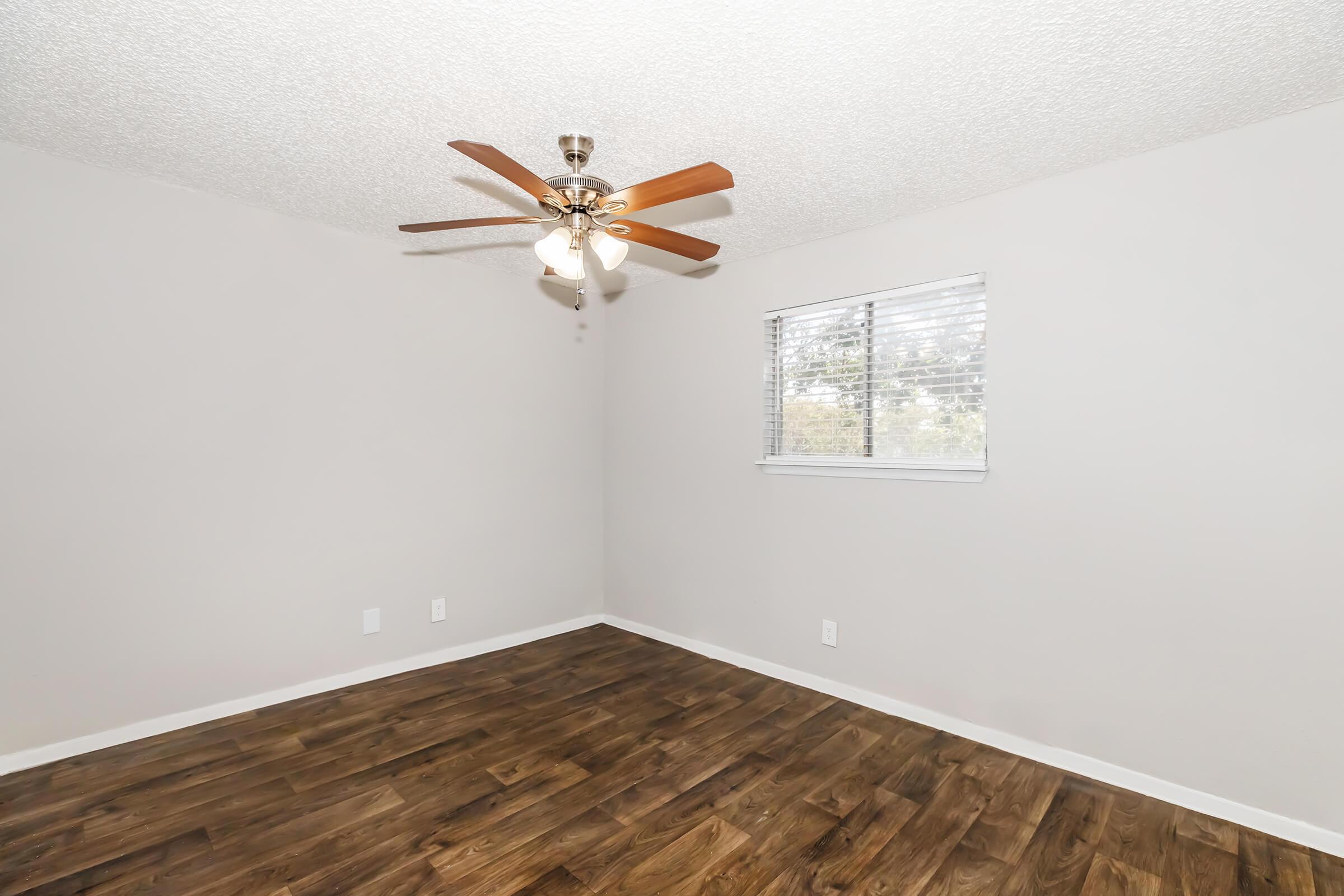 A spacious bedroom featuring light gray walls, a ceiling fan with wooden blades, and a large window with blinds allowing natural light. The floor is made of dark laminate wood. The room is unfurnished and has a clean, modern appearance.