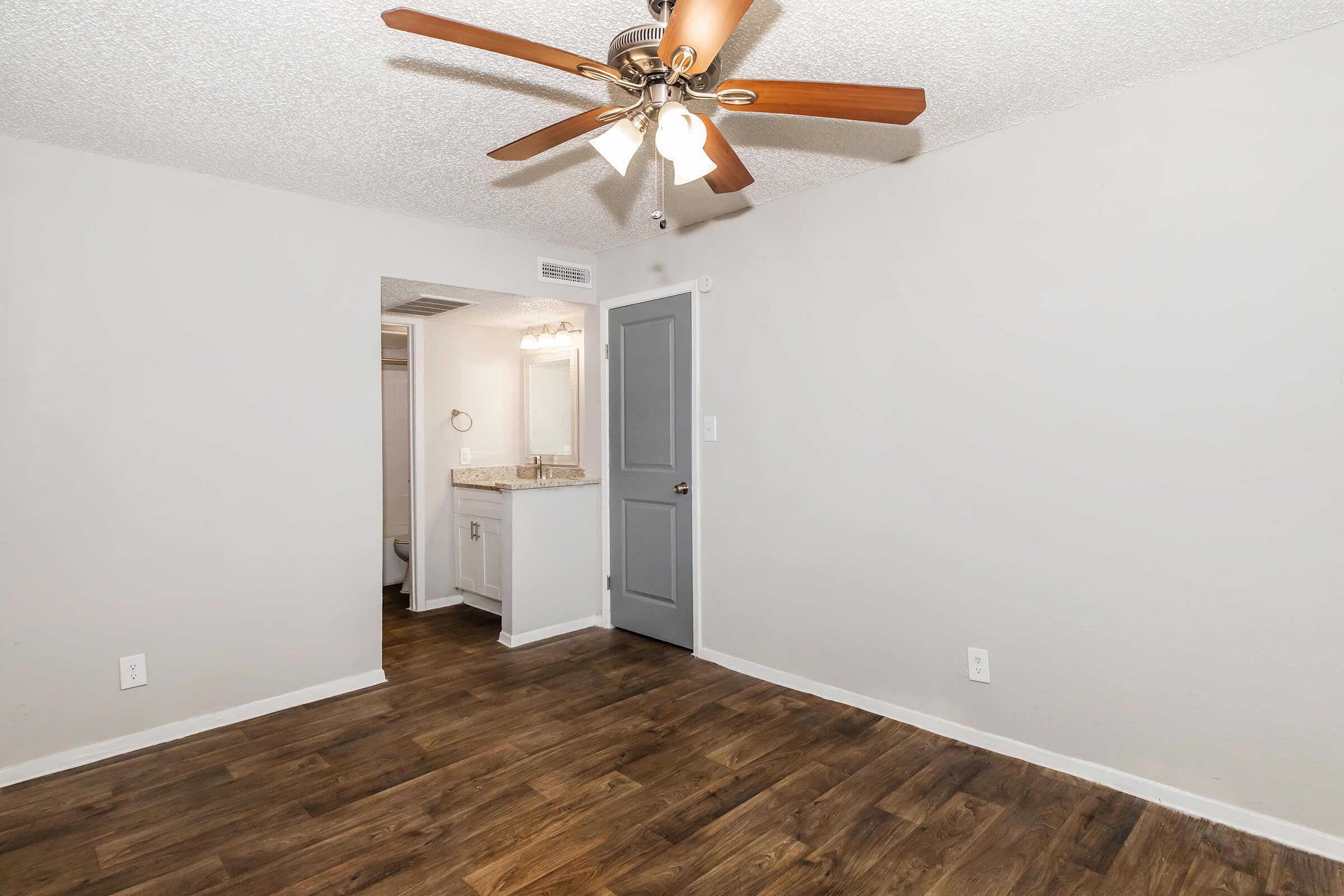 Interior view of a room featuring a ceiling fan with wooden blades, light-colored walls, and a wood-look floor. There's a doorway leading to what appears to be a bathroom, and a small vanity area with a mirror visible. The space is well-lit and can be used as a living area or bedroom.