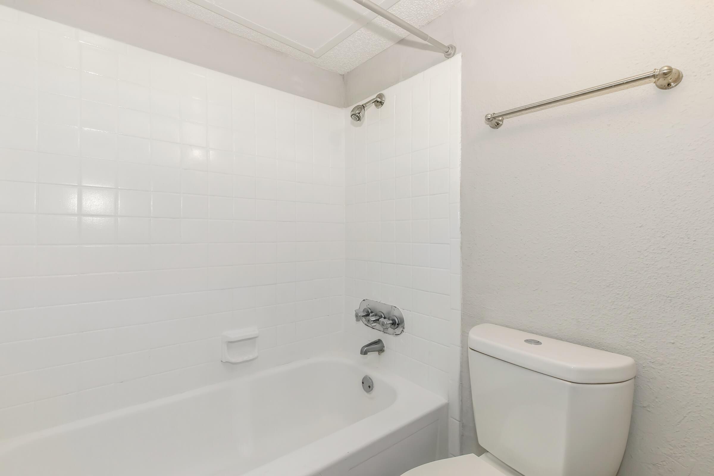 A clean, minimalist bathroom featuring a white bathtub and shower with a tiled wall. There is a modern toilet beside the bathtub, and a towel rack mounted on the wall. The overall aesthetic is bright and simple, with neutral colors and good lighting.