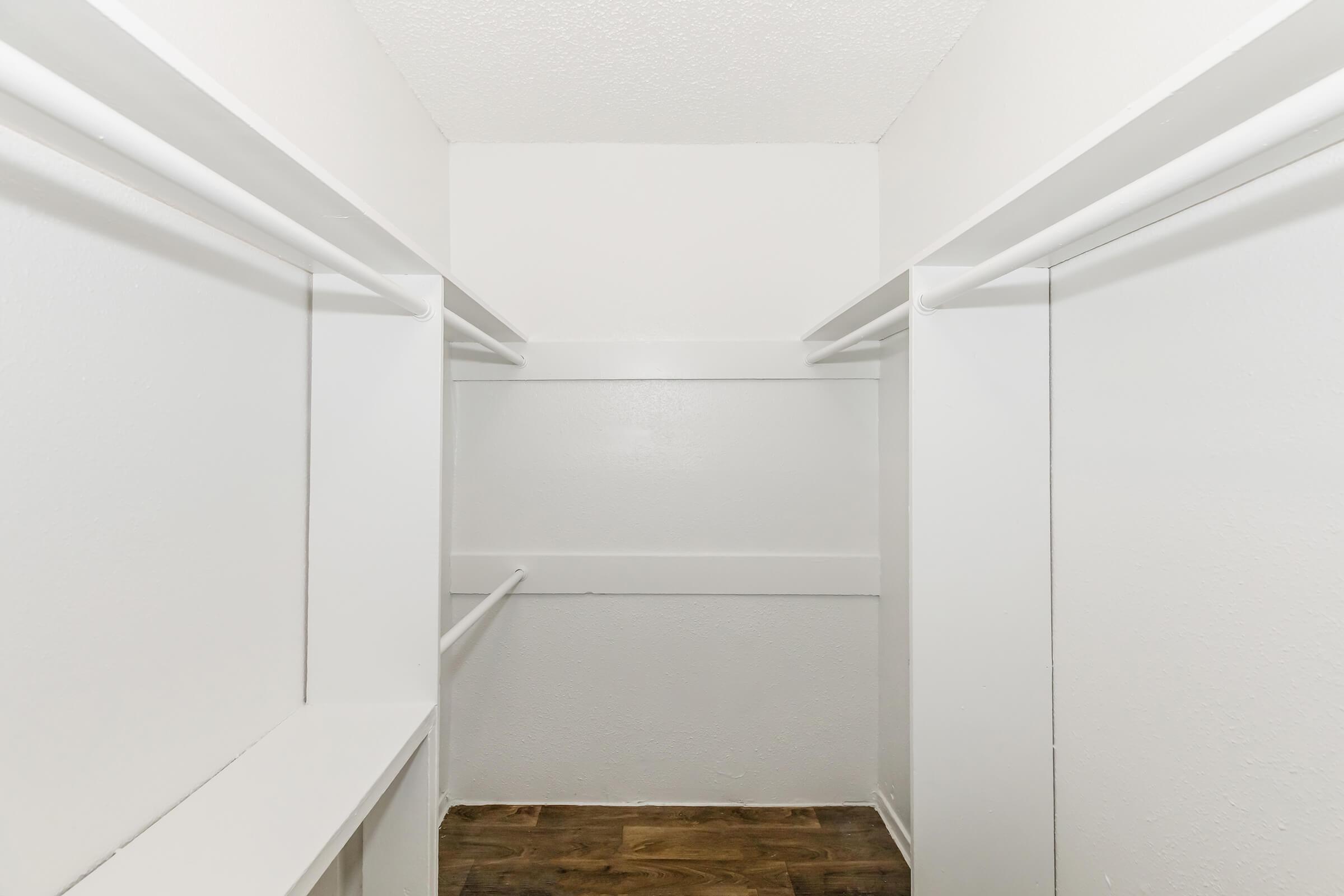 A spacious, empty closet featuring white walls and shelving on both sides. The floor is made of dark wood, providing a contrast to the bright interior. The closet has a simple, minimalist design with ample hanging space.