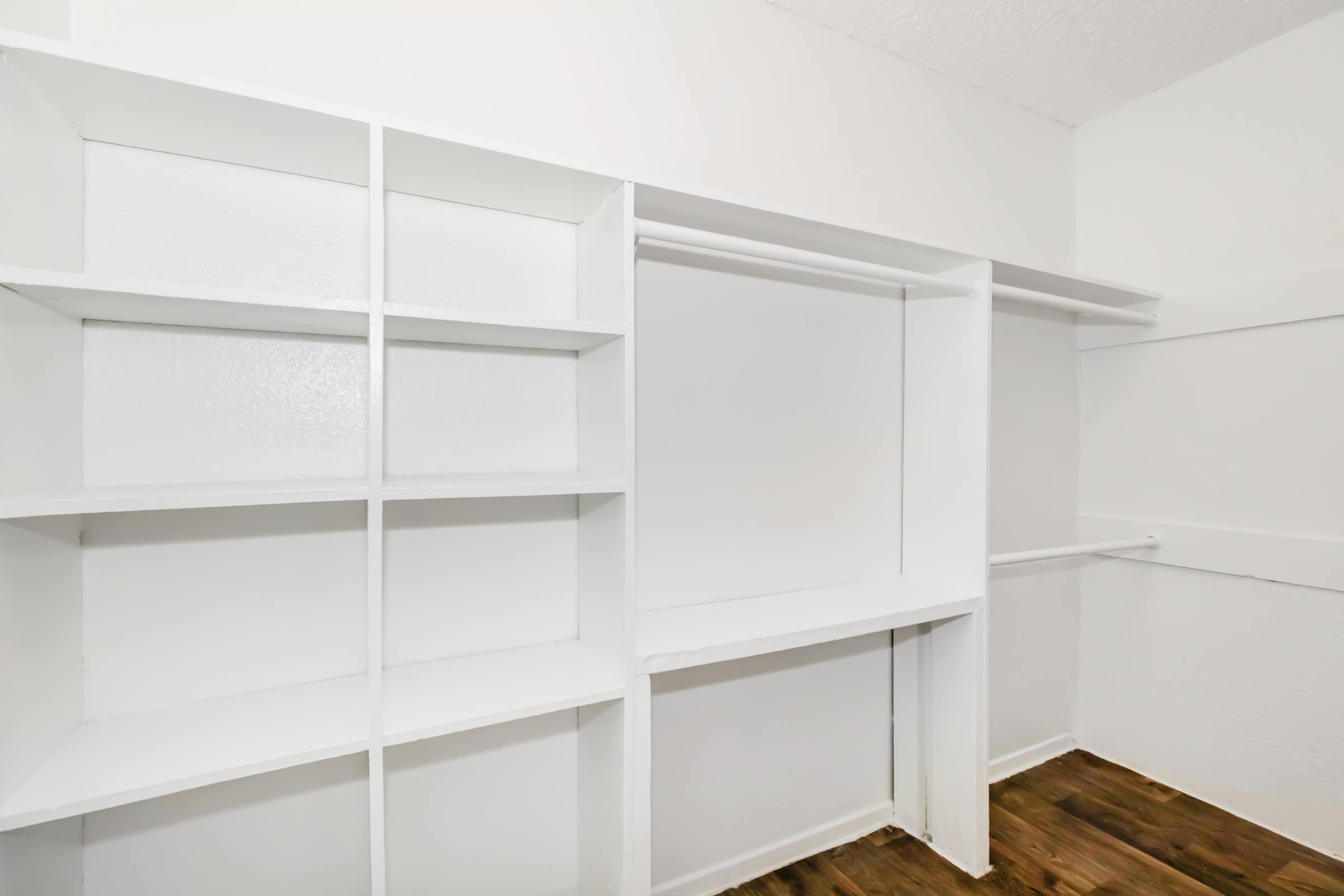 Empty closet space with white shelving units and a hanging rod. The walls are painted white, and the floor is wooden. Ideal for organizing and storing clothes or other items.