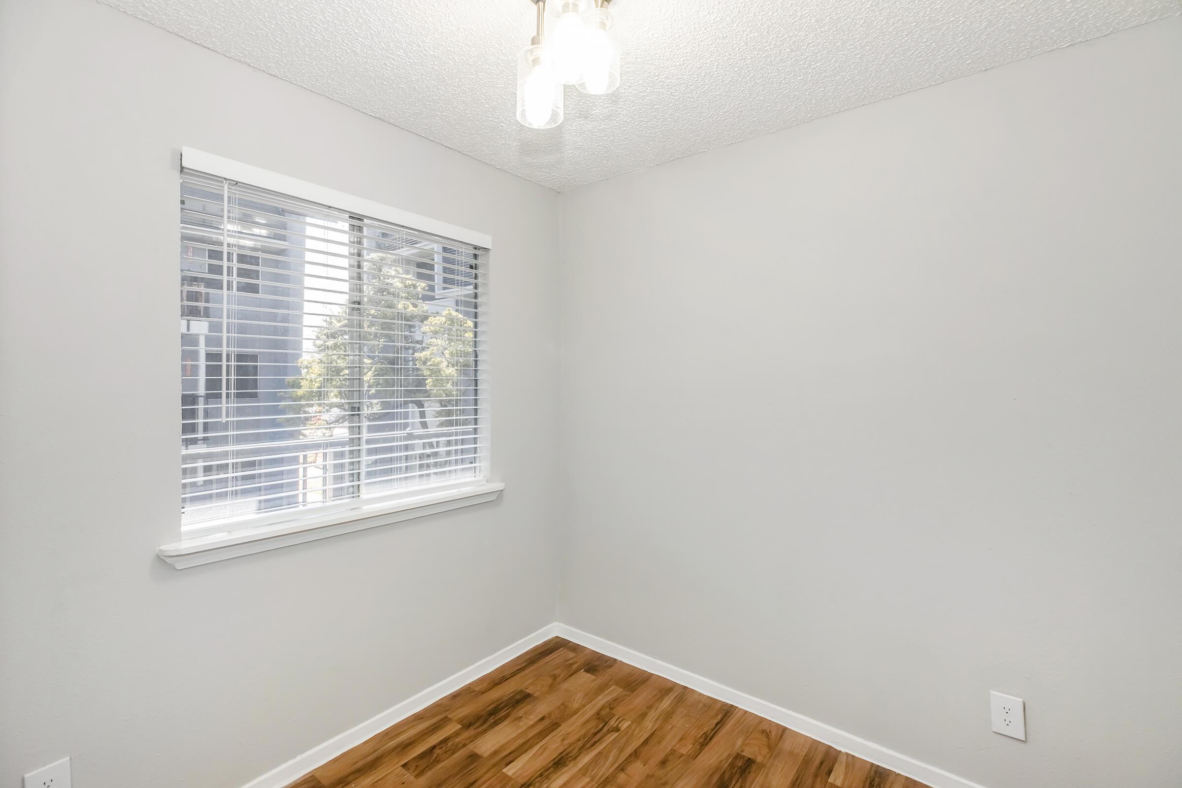 A small, empty room with light-colored walls and a wooden floor. A window with closed white blinds allows natural light to enter, and a simple light fixture hangs from the ceiling. The room appears clean and well-maintained, ready for decoration or furniture.