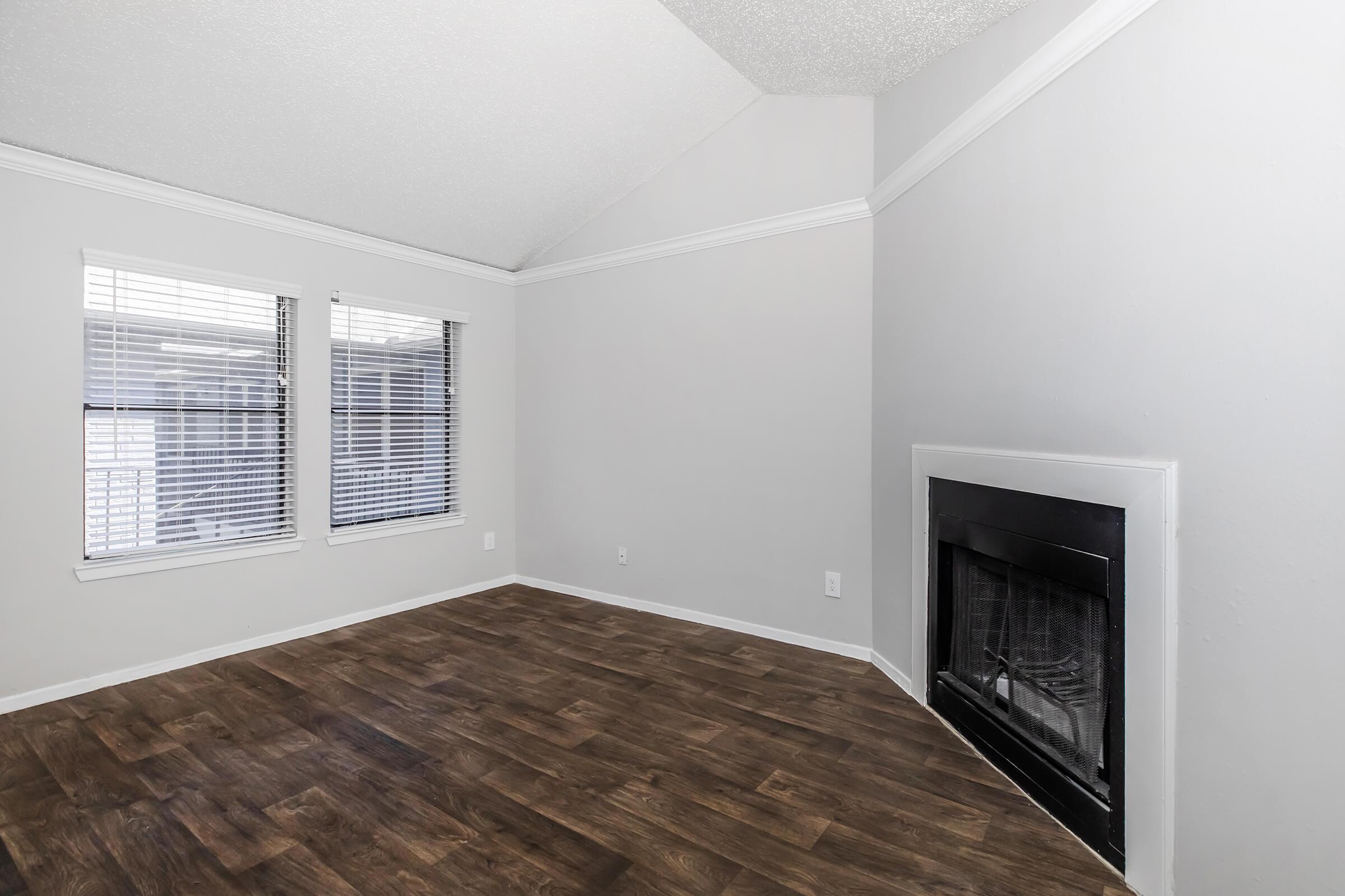 A spacious, empty room featuring a vaulted ceiling, light gray walls, and hardwood floors. There are two windows with white blinds allowing natural light. A black fireplace is positioned against one wall, contributing to the cozy ambiance.