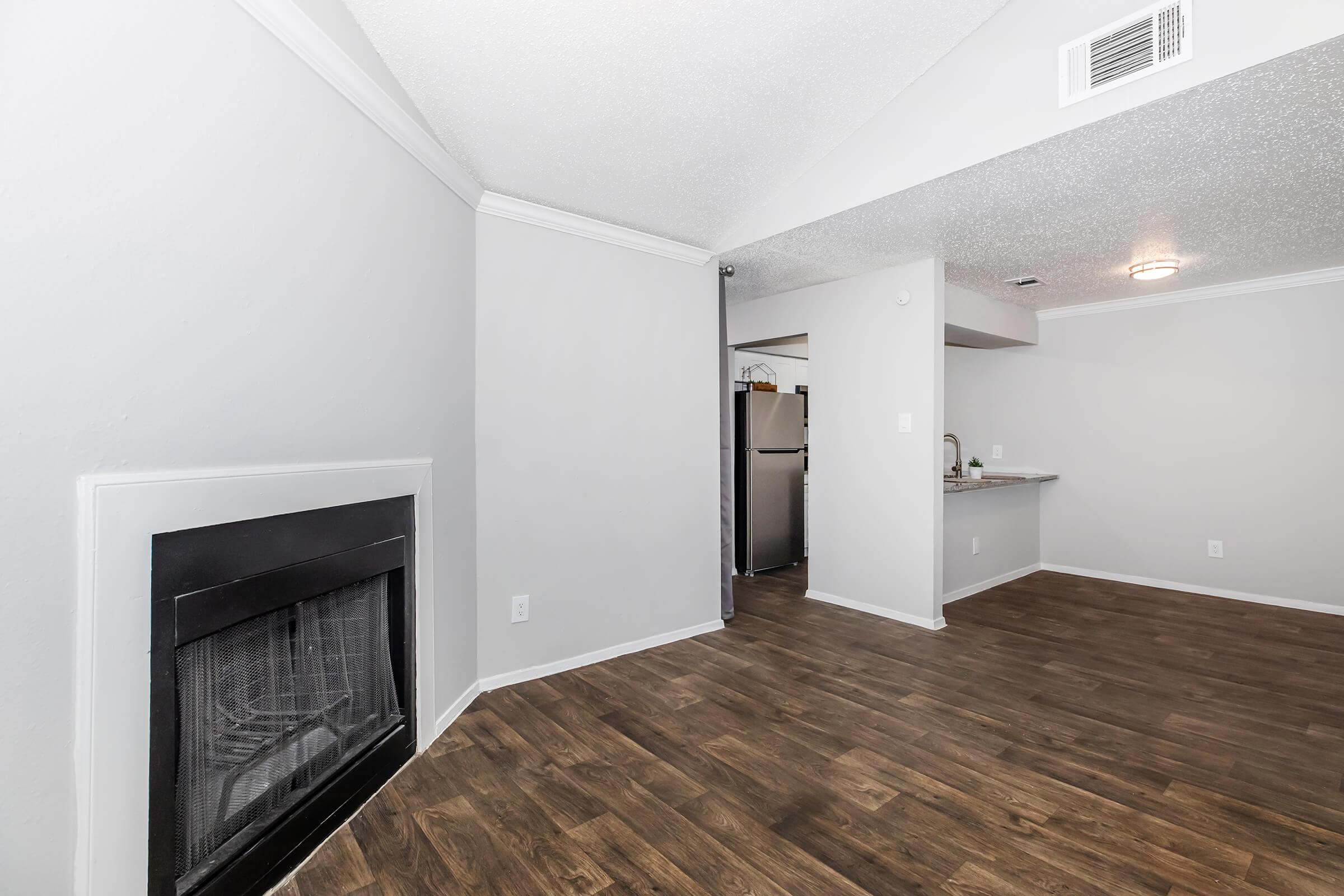 A living room with wood-style flooring, featuring a modern fireplace on the left. In the background, there's an open kitchen area with stainless steel appliances visible. The walls are painted in a light color, and there is a ceiling with crown molding, giving the space a contemporary feel.