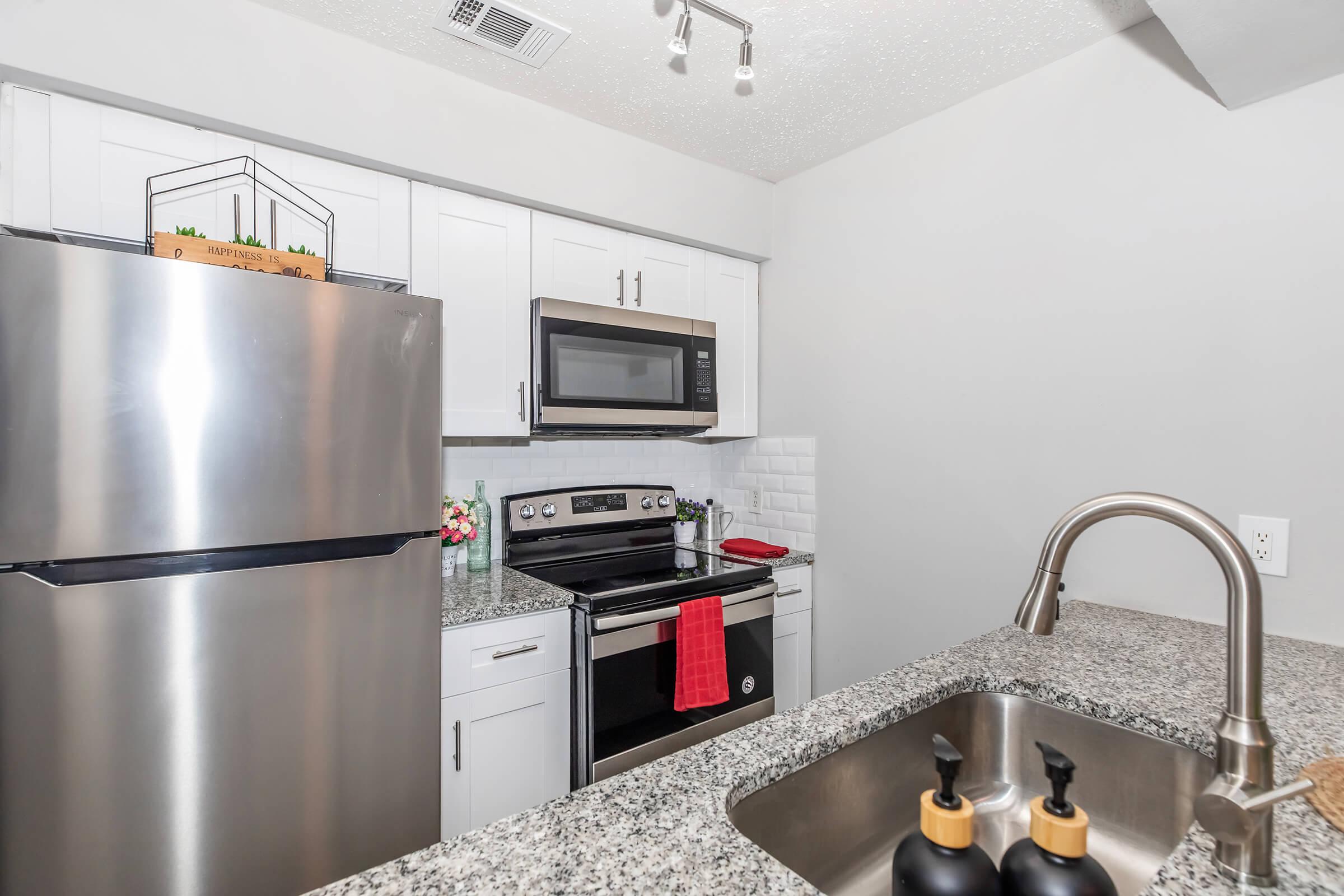 Modern kitchen with stainless steel appliances including a refrigerator and oven. The countertop features a granite surface with a sink and decorative items. A red kitchen towel hangs on the stove. White cabinets and a tiled backsplash create a bright, clean aesthetic.