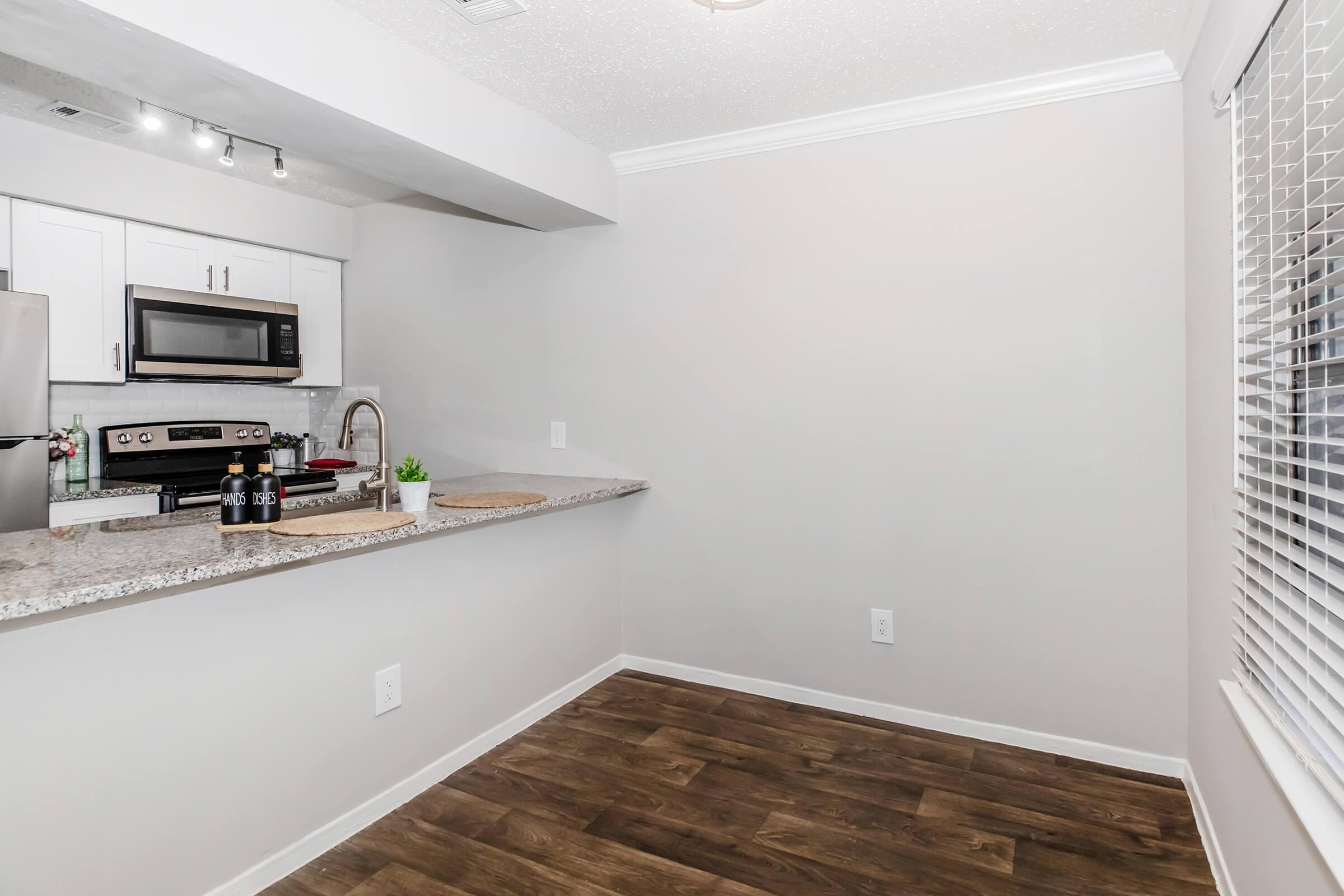 A modern kitchen with a granite countertop, stainless steel appliances, and a sink. There's a small bar area with bottles and a cutting board, and a light-colored, partially open space with wood flooring. A window with blinds allows natural light into the room.