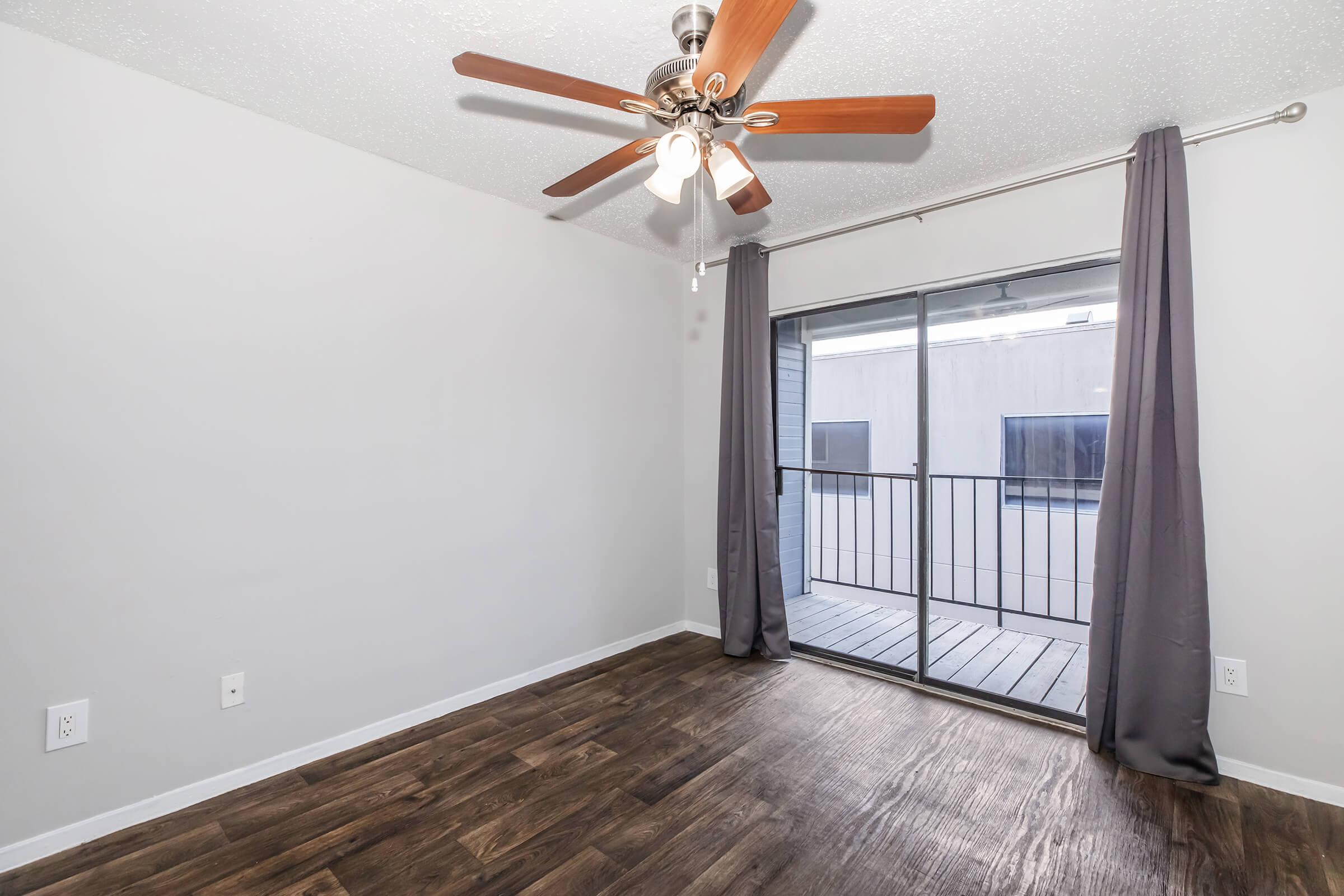 A spacious room with hardwood-style flooring, featuring a ceiling fan with wooden blades, light-colored walls, and a sliding glass door leading to a small balcony. Sheer curtains frame the door, allowing natural light to enter. The overall atmosphere is bright and inviting.