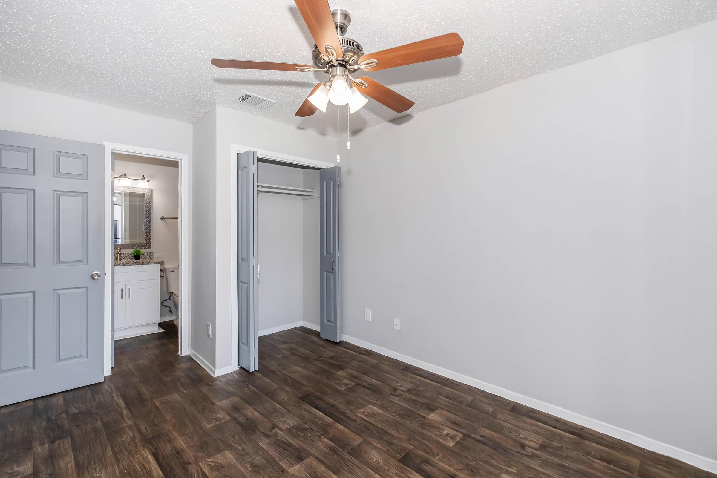 A spacious, well-lit room featuring a ceiling fan with wooden blades, light gray walls, and a dark wood floor. There are two open doors leading to a closet and an adjoining bathroom visible in the background. The overall design is modern and clean.