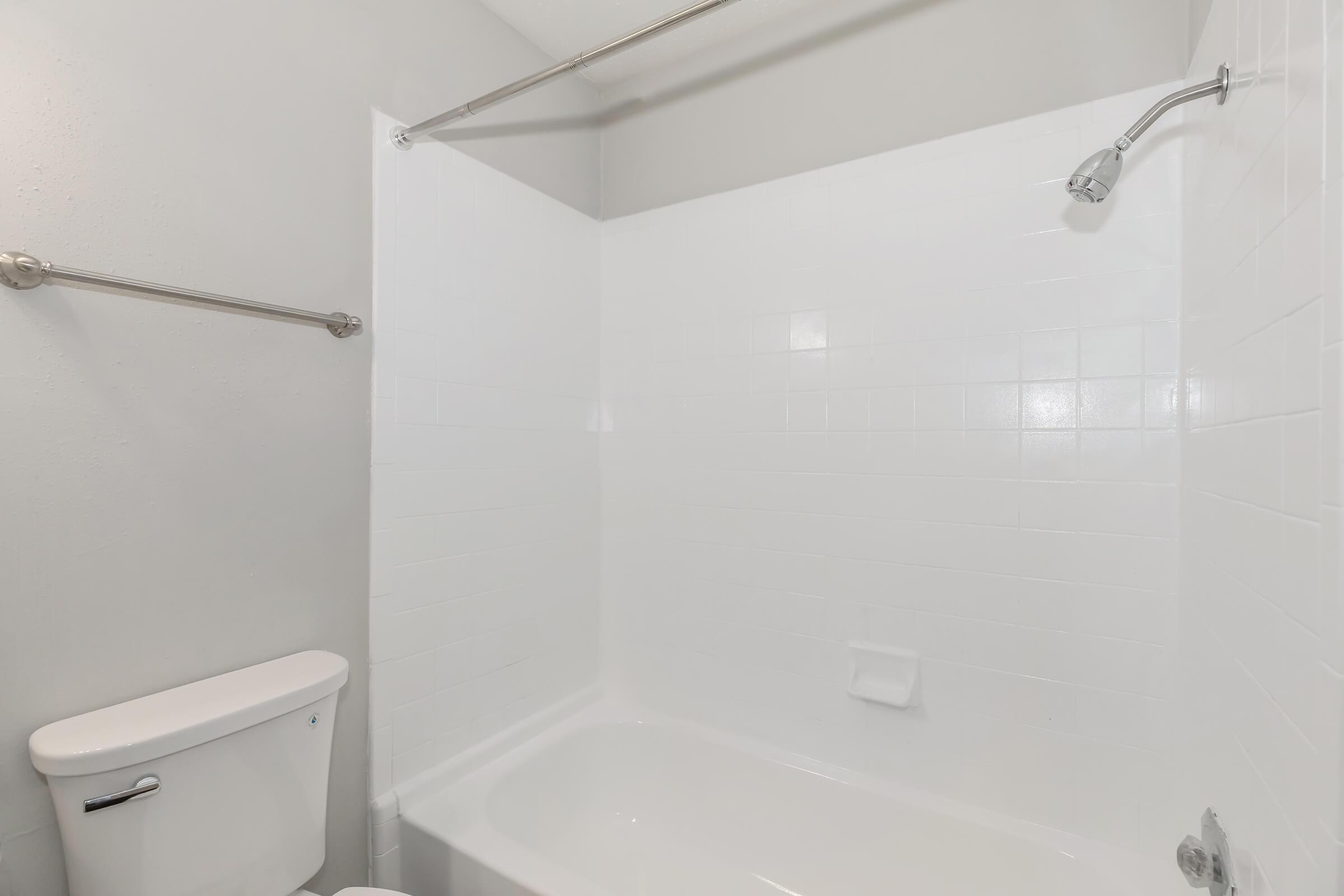 A clean bathroom featuring a white bathtub, a showerhead above, and a toilet beside it. The walls are painted light gray, and the tile is white, creating a bright and simple aesthetic.