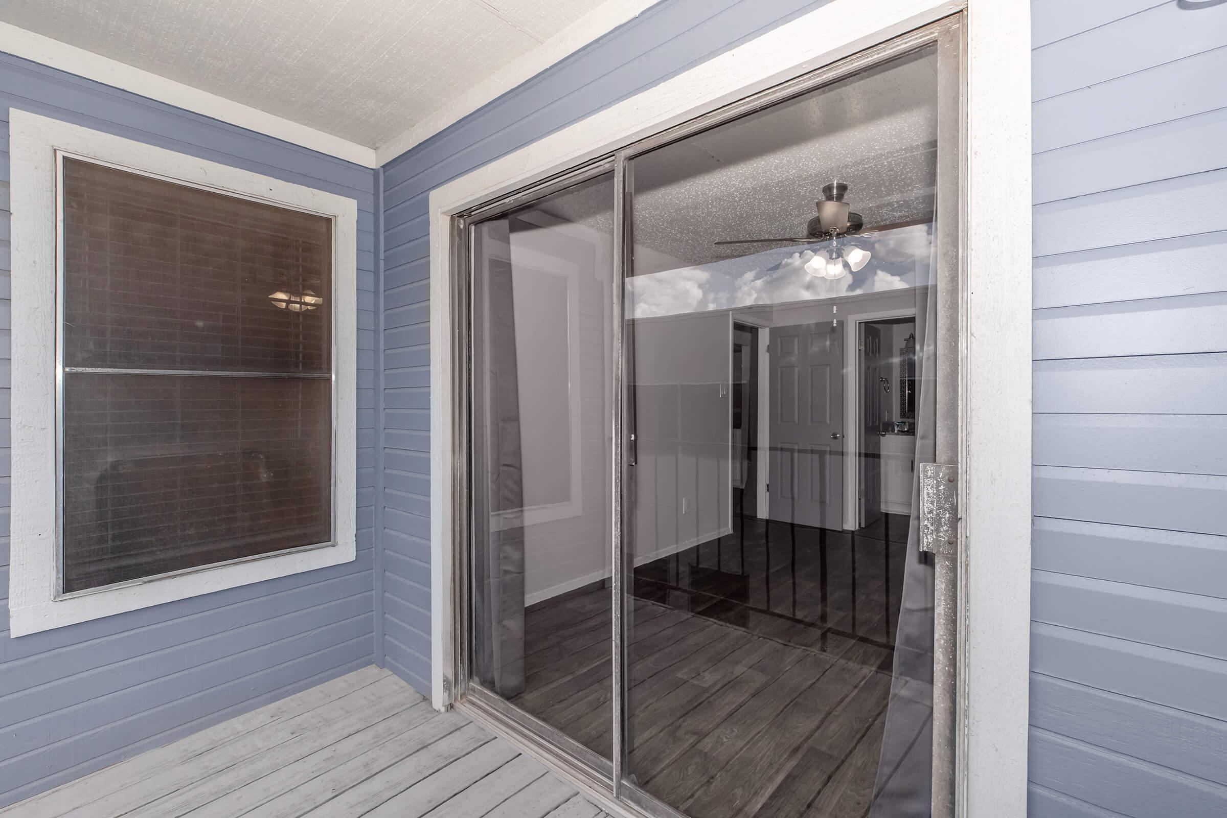 View of a sliding glass door leading from a patio to a room with light-colored walls and a ceiling fan. The exterior features blue siding, and there is a large window on the left. The interior floor is tiled, creating a modern and open atmosphere.