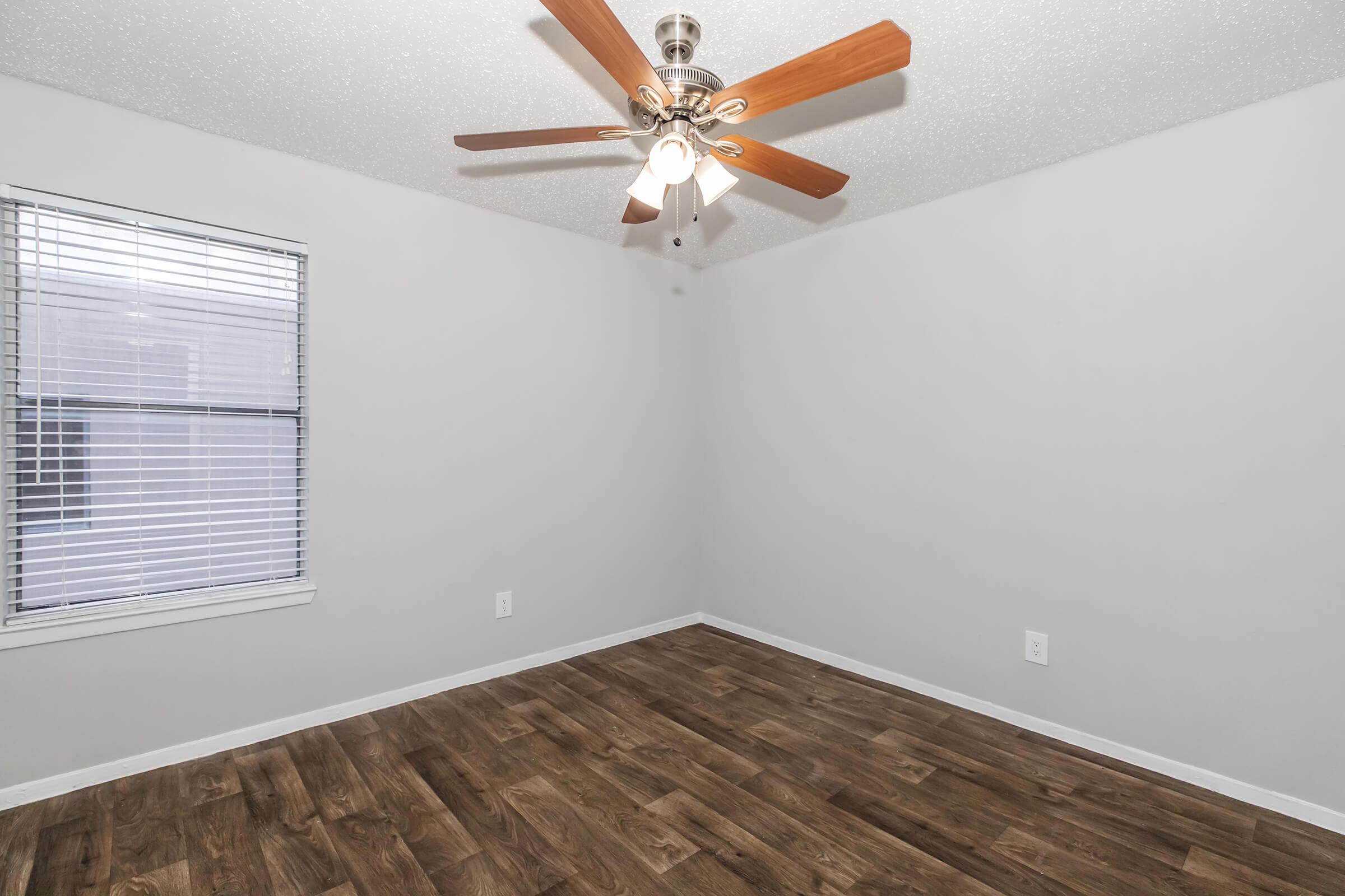 A spacious, empty room featuring a ceiling fan with wooden blades, light-colored walls, and a window with blinds. The floor is covered with brown wooden plank-like flooring, giving a warm and inviting appearance.