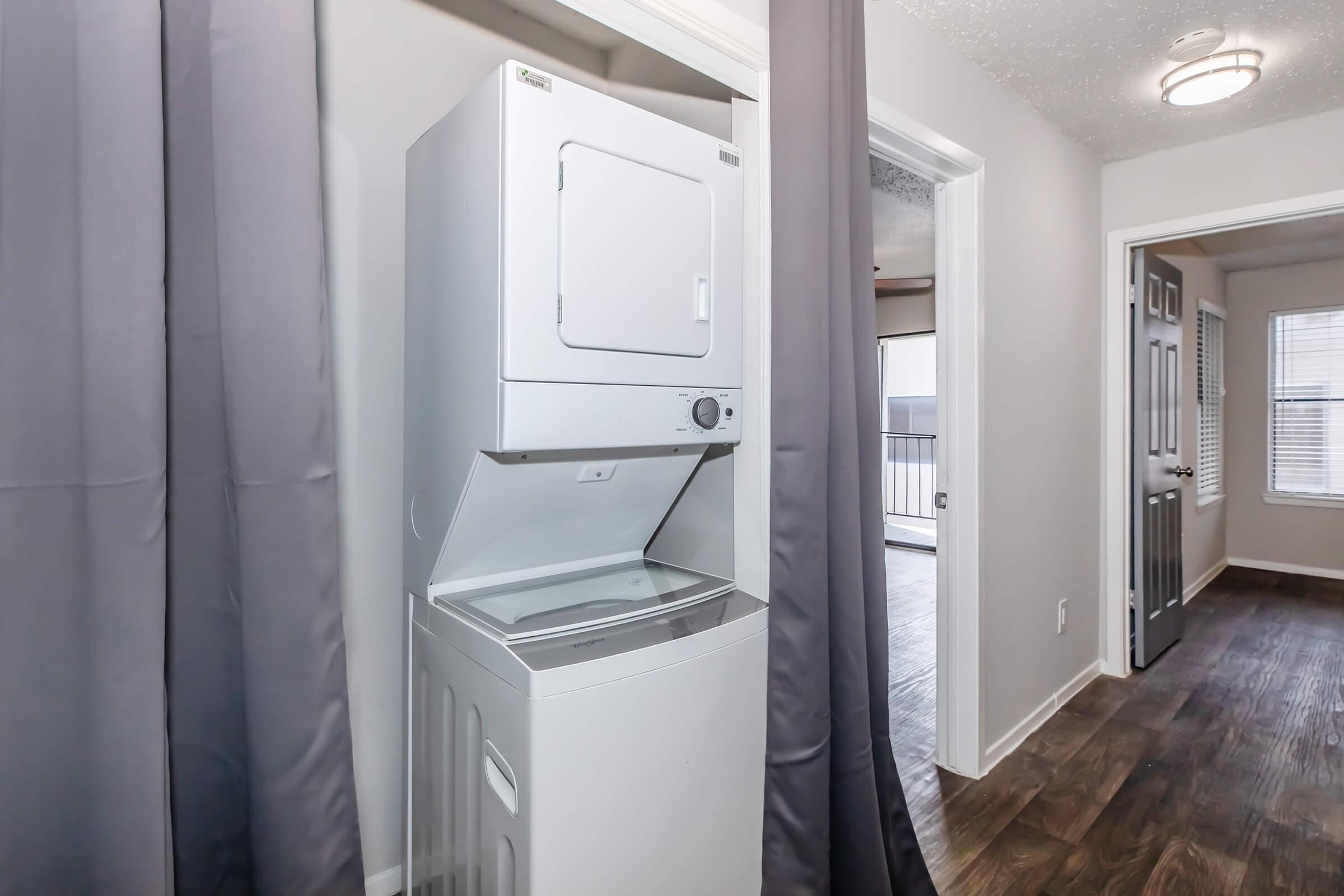 A stacked washer and dryer unit situated in a narrow hallway, with gray curtains partially covering the area. The hall leads to a room with a door, and there is a light fixture on the ceiling. The flooring appears to be wood laminate, and there are windows visible in the adjacent room.