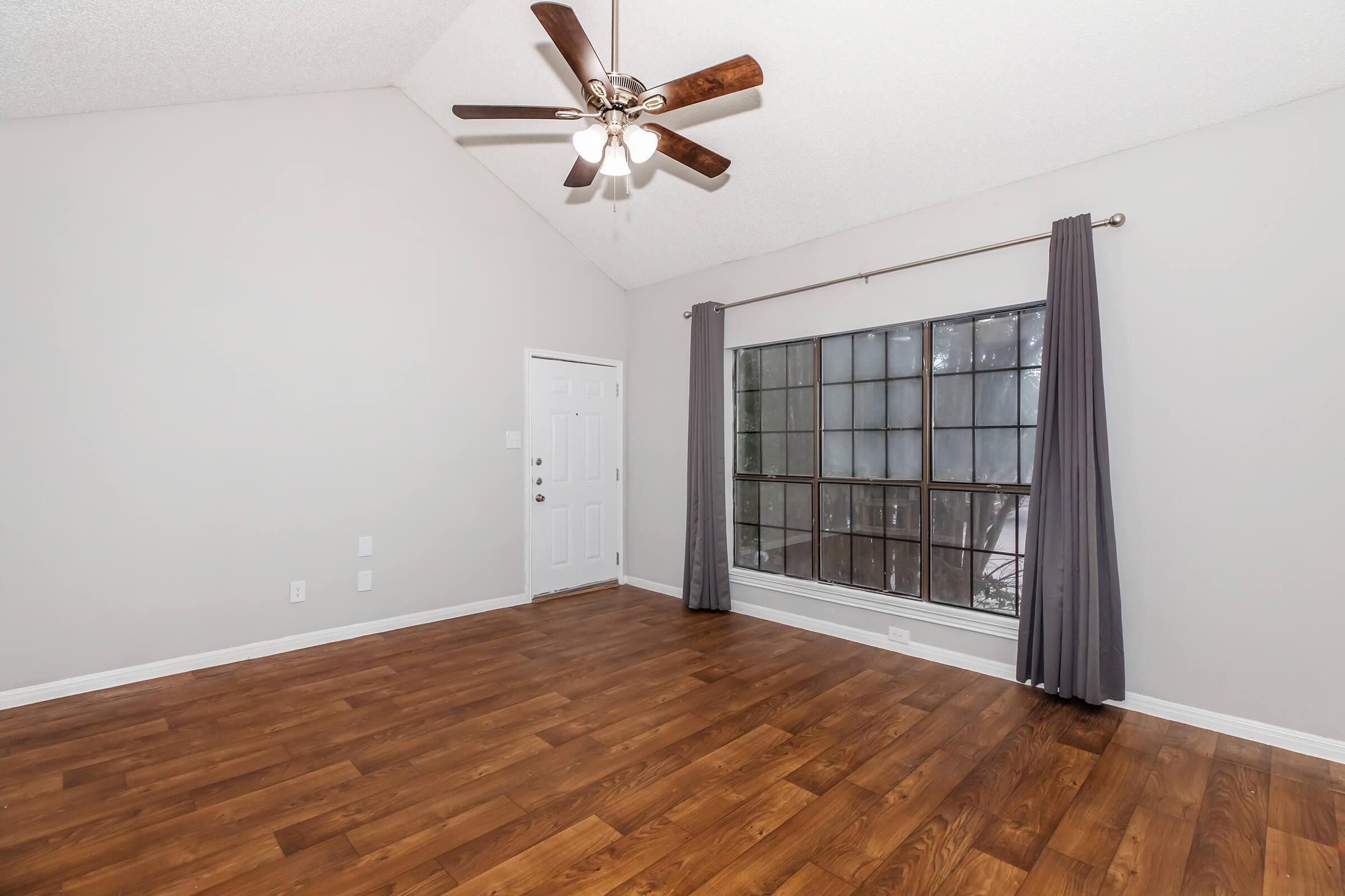 A spacious, well-lit room featuring a ceiling fan, hardwood flooring, and a large window with sheer curtains. The walls are painted in a light color, and there is a front door visible. The room has a high ceiling, giving it an airy feel.