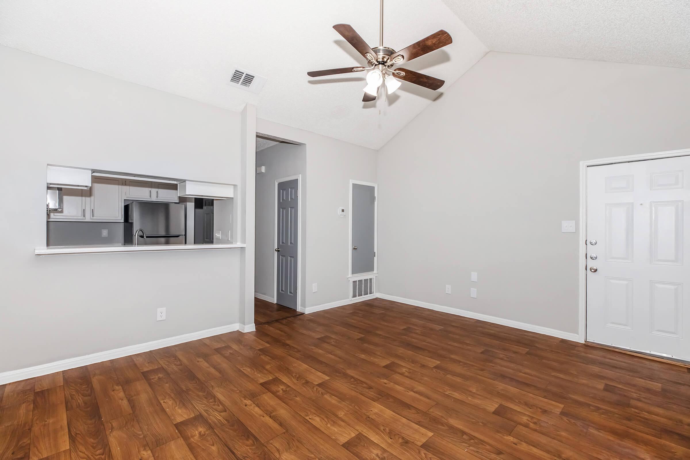 A spacious living room featuring hardwood floors, a ceiling fan, and a vaulted ceiling. It includes a doorway leading to another room and an open view towards a kitchen area with a breakfast bar. The walls are painted in light neutral colors, enhancing the airy feel of the space.
