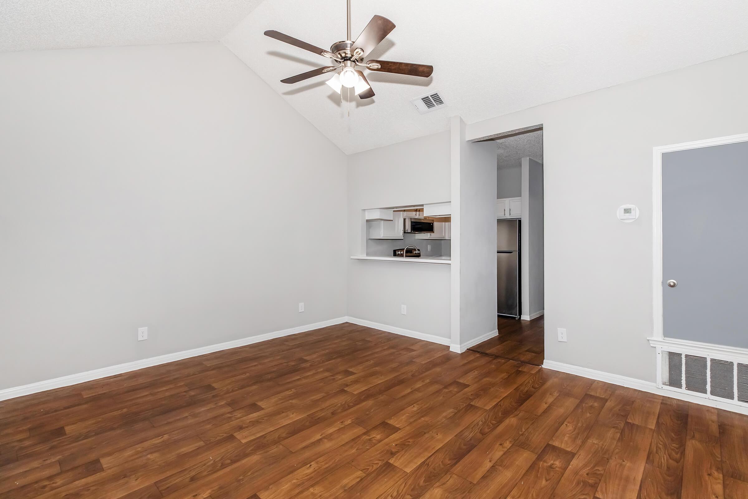 A spacious living area with a high ceiling, featuring a ceiling fan and hardwood flooring. A small kitchen with a pass-through is visible in the background, alongside a doorway leading to another room. The walls are painted in neutral tones, enhancing the bright and open feel of the space.