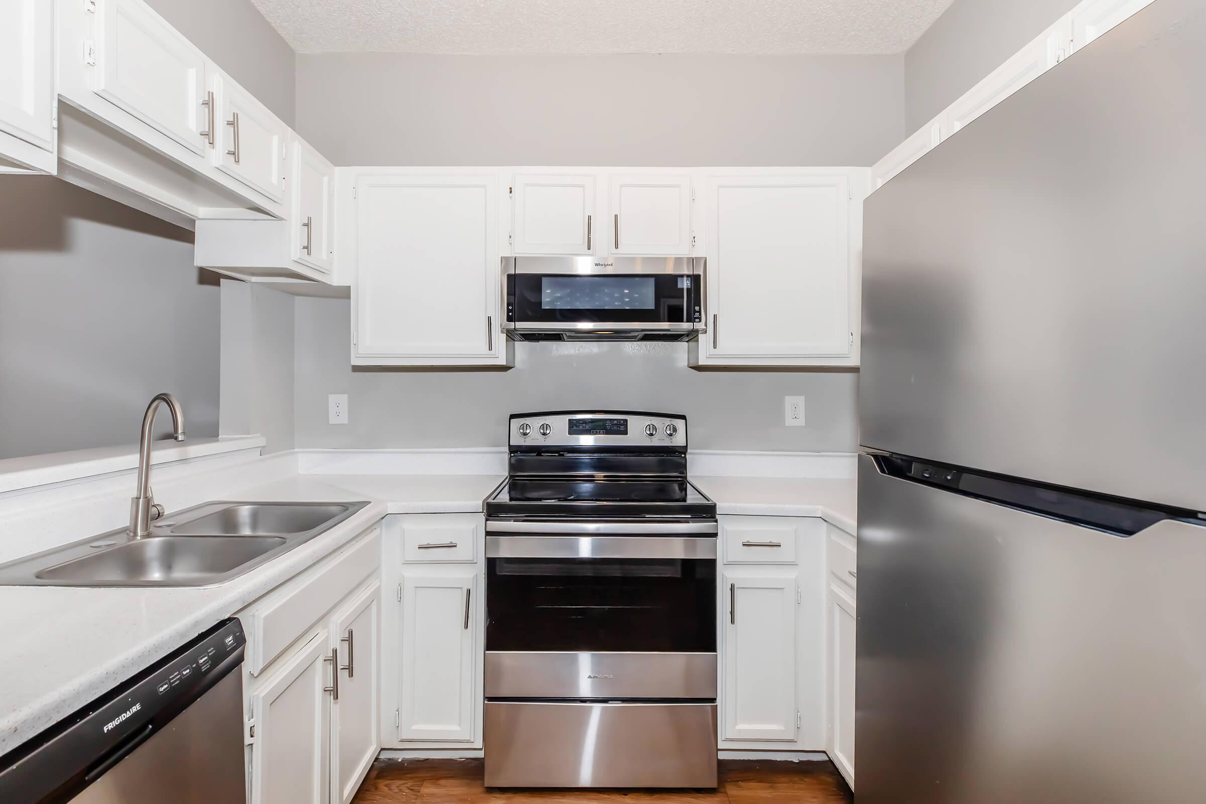 Modern kitchen featuring white cabinetry, stainless steel appliances including a microwave, electric stove, and refrigerator. The countertops are light-colored and there are double sinks. The walls are painted in a soft gray, creating a clean and contemporary look.