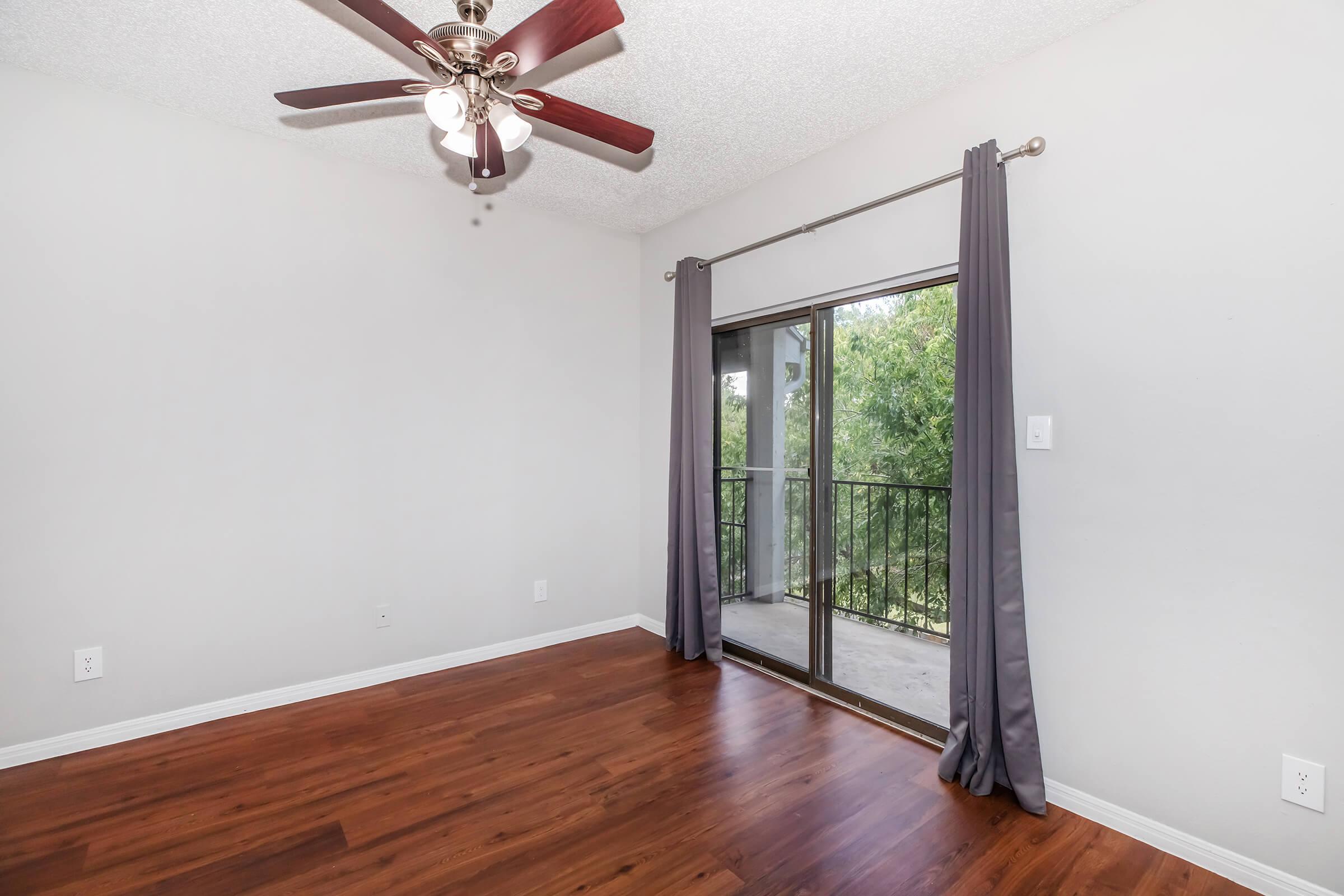 A well-lit room featuring a ceiling fan with wooden blades, neutral gray walls, and dark hardwood flooring. A set of sliding glass doors leads to a small balcony with outdoor views, framed by floor-length gray curtains. The space is empty, providing a clean and airy feel.