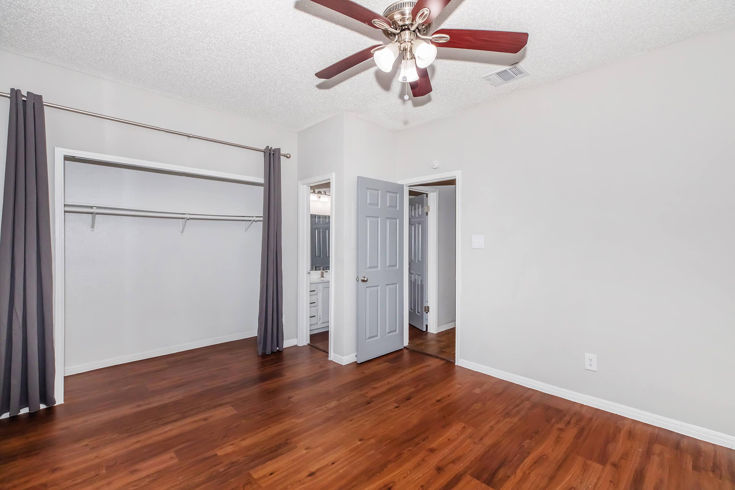 A spacious room with hardwood flooring, featuring a ceiling fan with light fixture. One wall has a closet with hanging rods, and there's a bathroom door on the right. The walls are painted in a light color, and there are two curtain panels on the left side of the room.