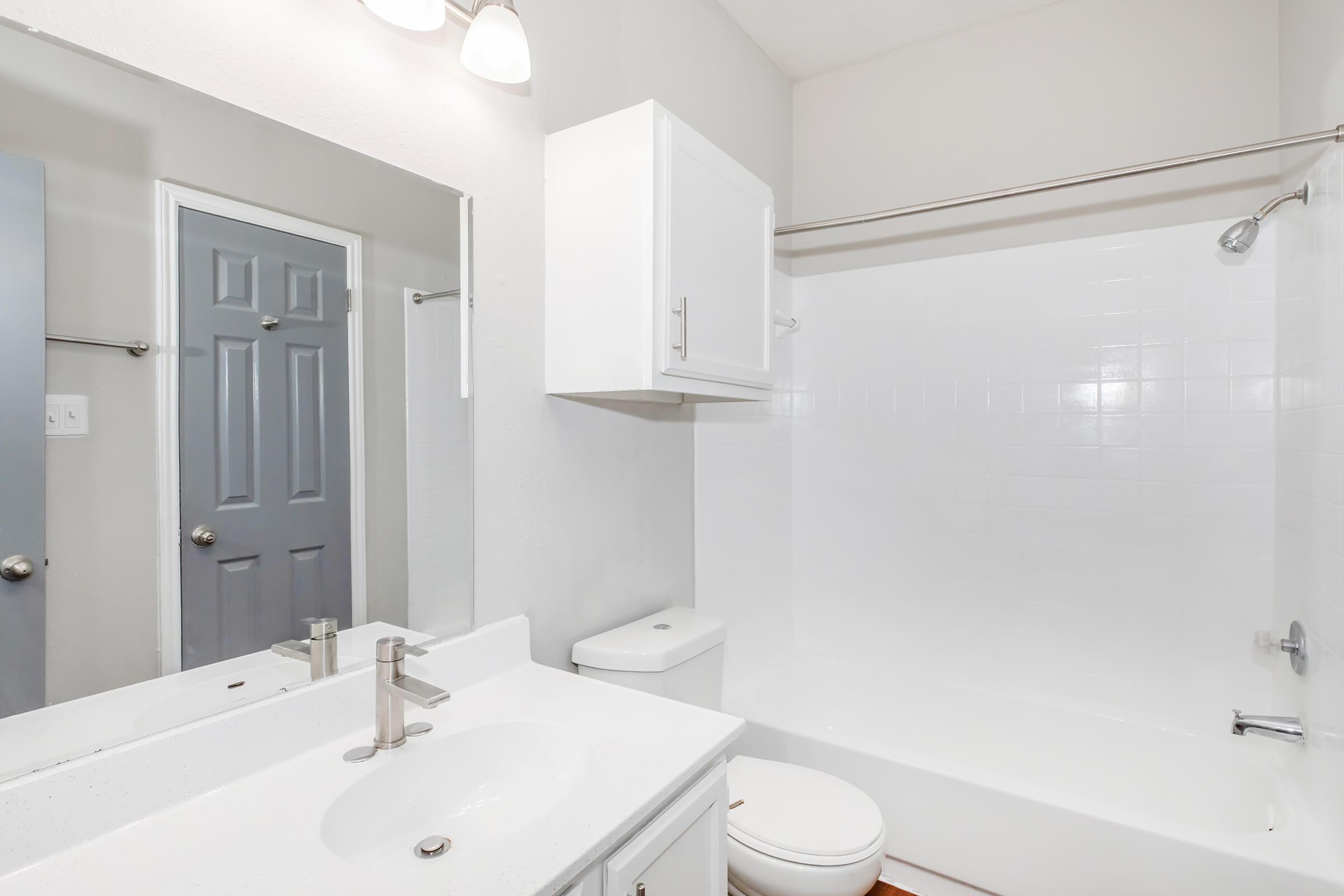 A clean and modern bathroom featuring a white bathtub, toilet, and sink with a sleek countertop. A large mirror above the sink reflects the bright space, while a gray door provides access to another room. The walls are painted a light color, enhancing the overall brightness of the area.