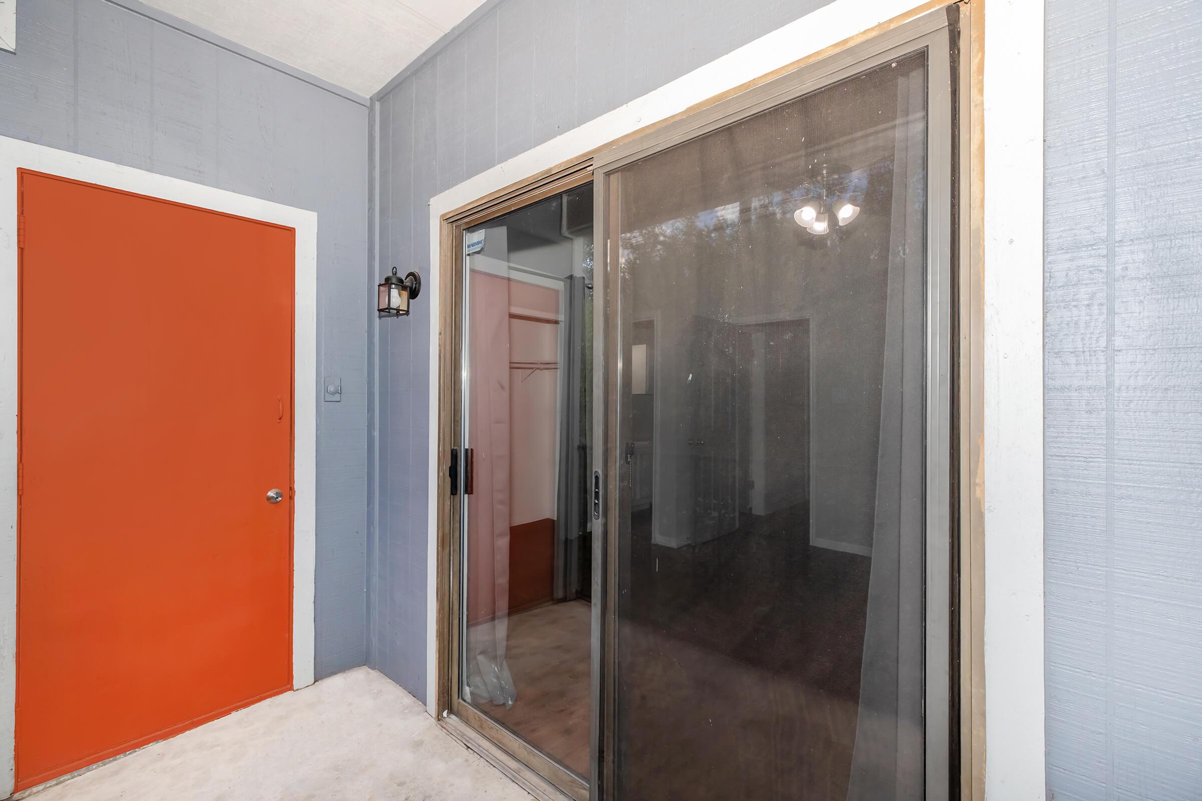 A porch area with a sliding glass door leading into a room. The walls are painted gray, and there is an orange door to the left. Inside, a soft pink wall can be seen through the glass. The space is well-lit, with a light fixture visible above. The flooring appears to be light-colored, adding to the open feel.