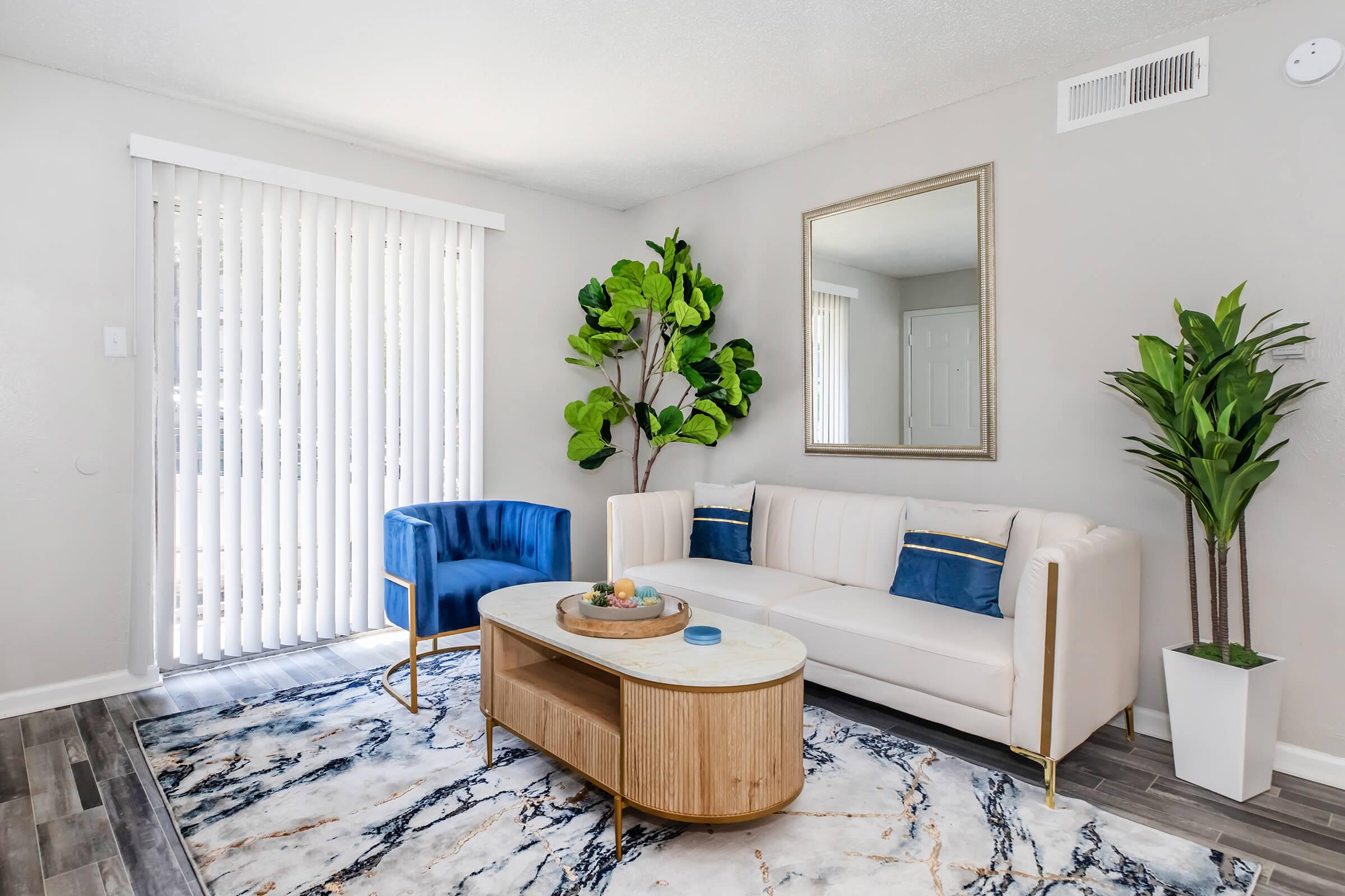 A modern living room featuring a white sectional sofa with blue and gold accents, a round wooden coffee table with decorative items, and two indoor plants. There is a large mirror on the wall and sliding glass doors covered with vertical blinds, allowing natural light into the space.