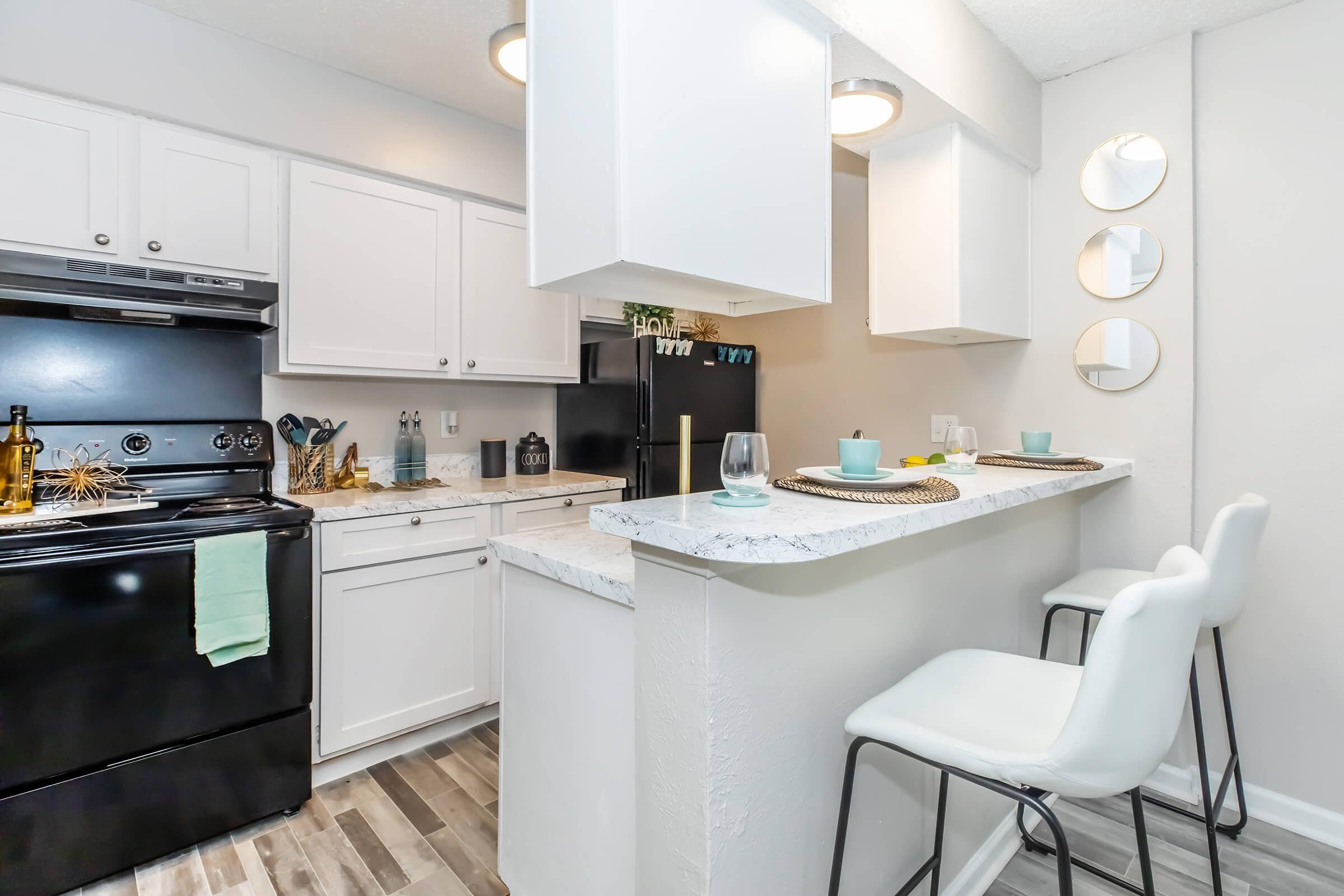 A modern kitchen with white cabinets and a black stove. The countertop features decorative items and two light blue bowls. In the foreground, two white bar stools are positioned at a small dining area. The wall is adorned with three round mirrors, adding a contemporary touch to the space. Bright lighting enhances the overall ambiance.