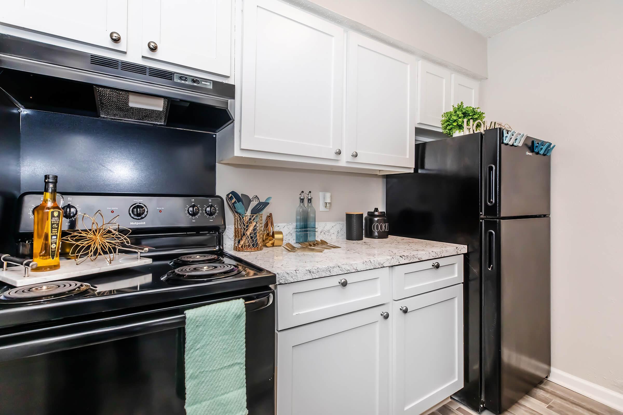 A modern kitchen featuring black appliances including a stove and refrigerator, white cabinets, and a granite countertop. The countertop has decorative items and kitchen utensils. A green towel is hanging by the stove, adding a pop of color to the clean and organized space.