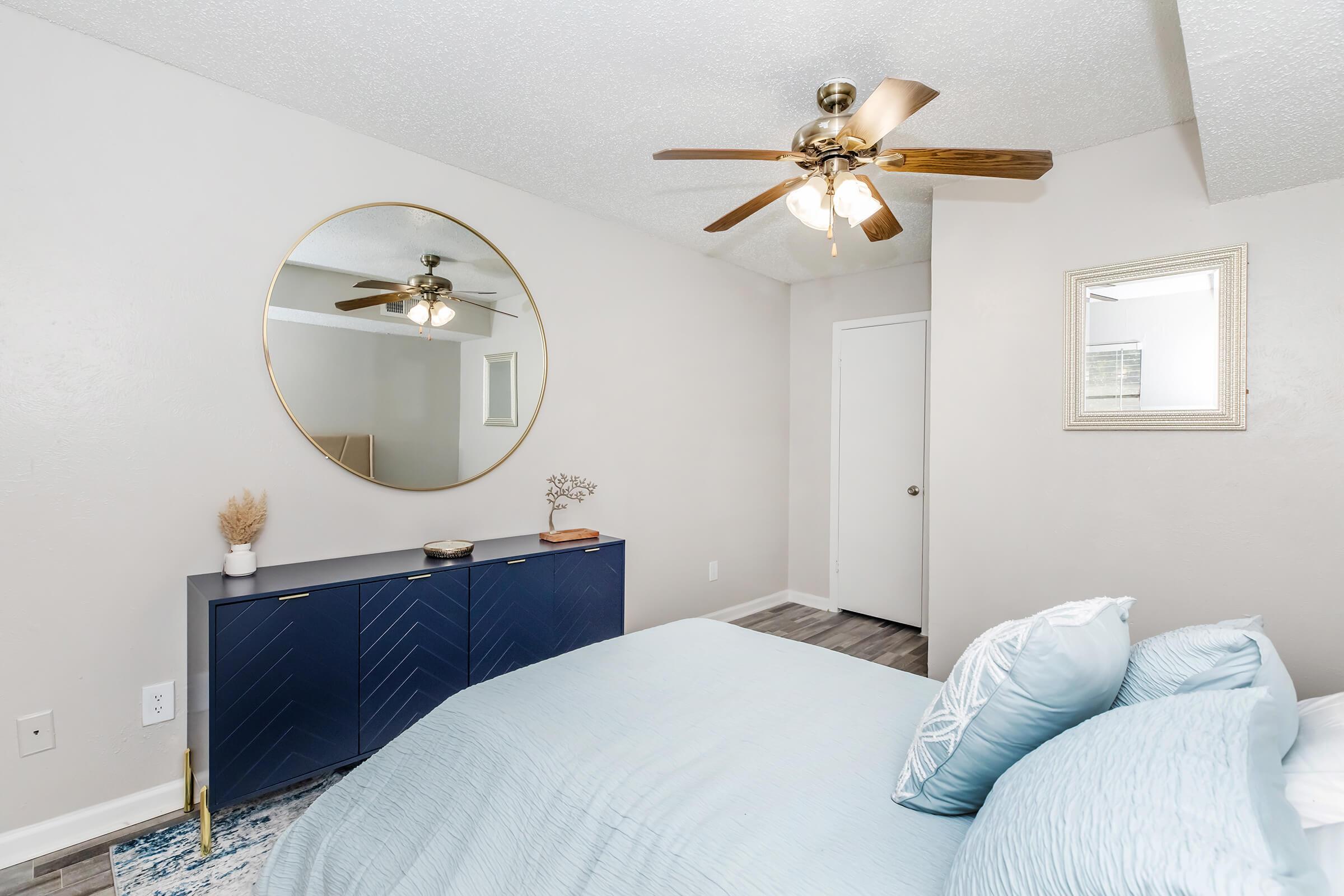 A cozy bedroom featuring a queen-sized bed with light blue bedding, a round mirror on the wall, and a modern ceiling fan. There is a dark blue dresser with decorative items, and the room has neutral-colored walls and flooring, creating a calming atmosphere. A door leads to another room.