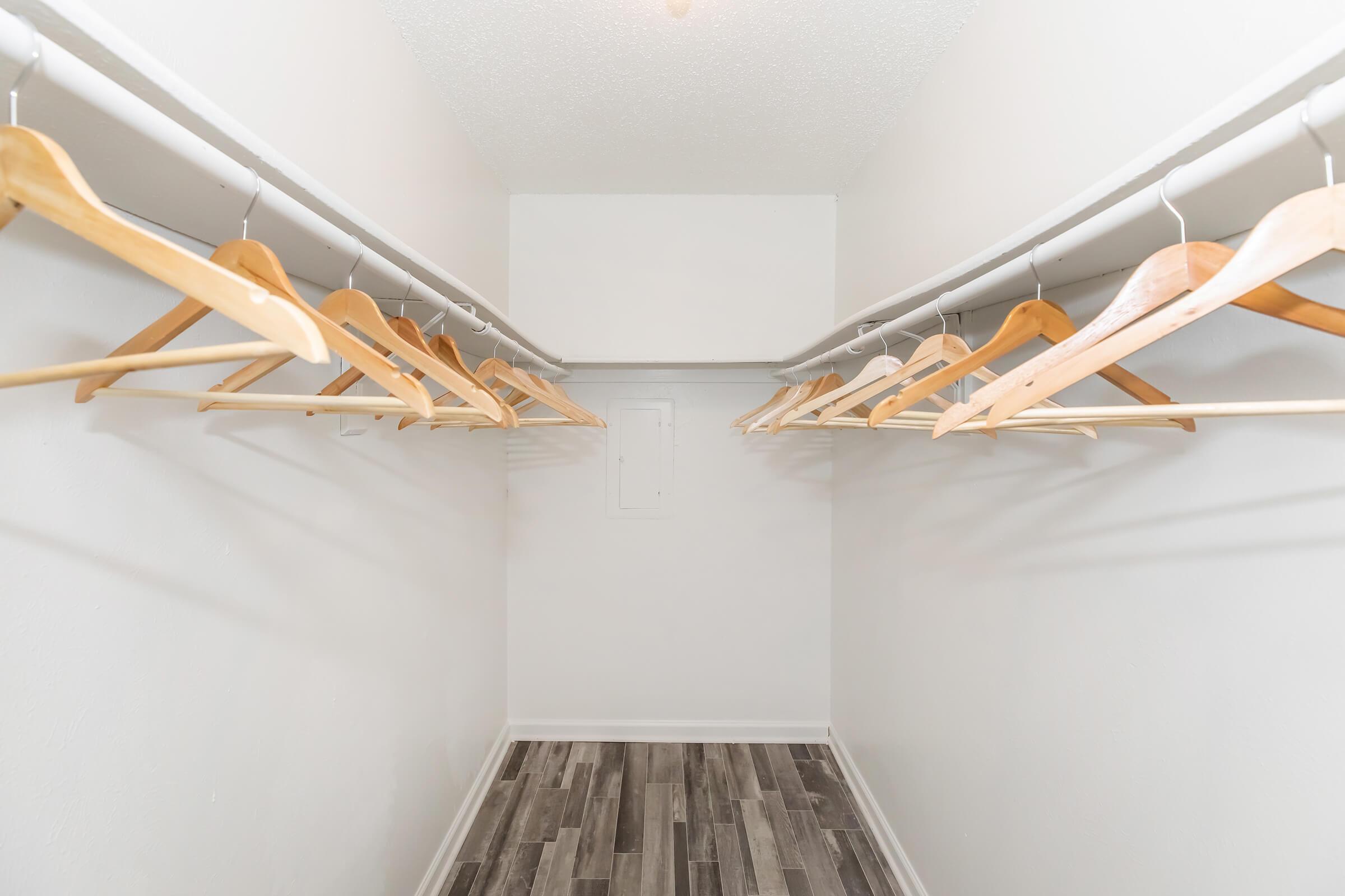 A spacious, empty walk-in closet featuring white walls and a gray wooden floor. Several wooden hangers are neatly arranged on two rods along the sides, highlighting the ample storage space available. Natural light may be coming from an overhead fixture.